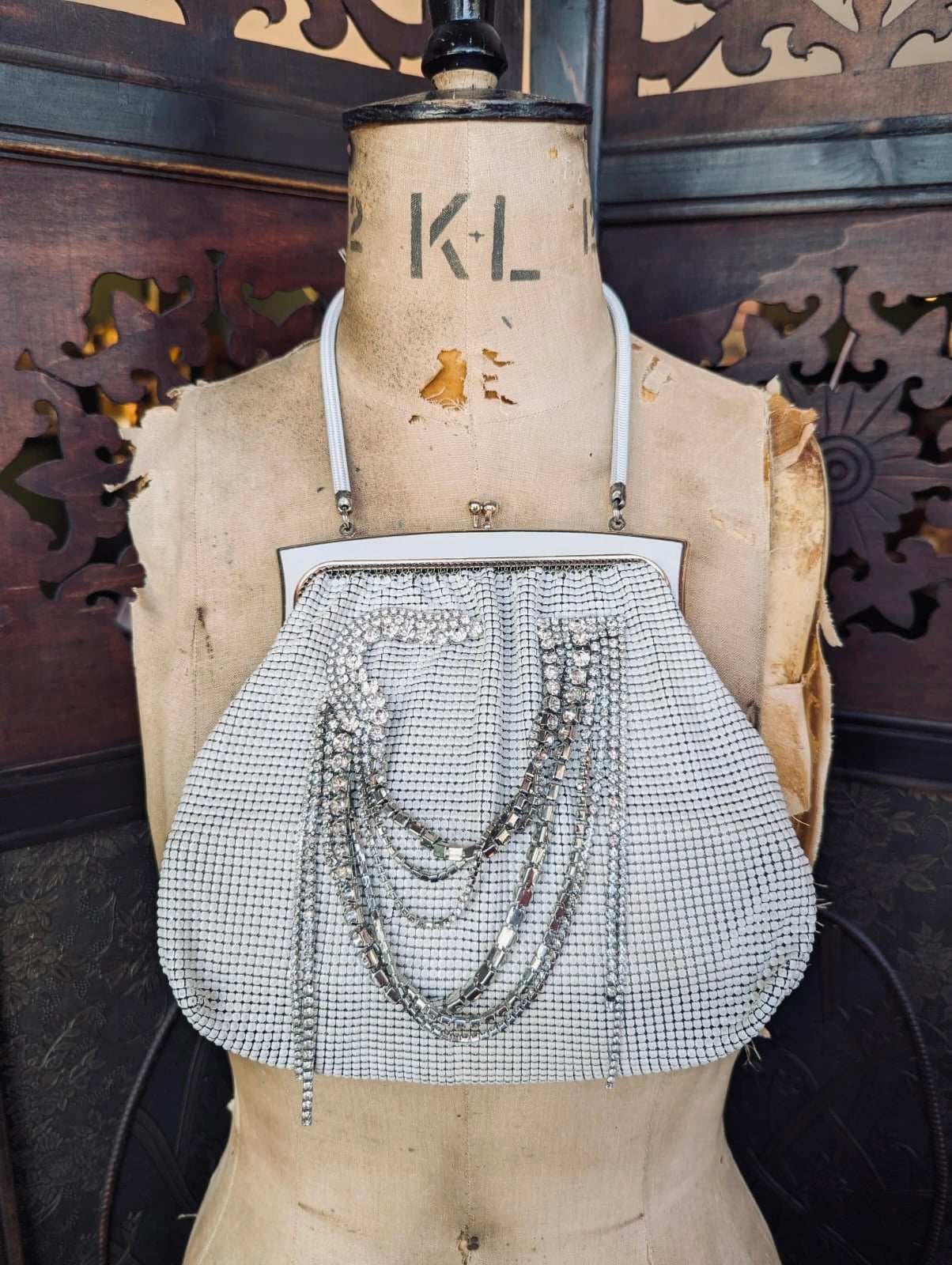 A Broochella Maison Broche l'orchidee vintage white glomesh clutch purse with a white floral brooch and white shoulder straps, displayed on a tan fabric mannequin torso against a dark carved wooden background.