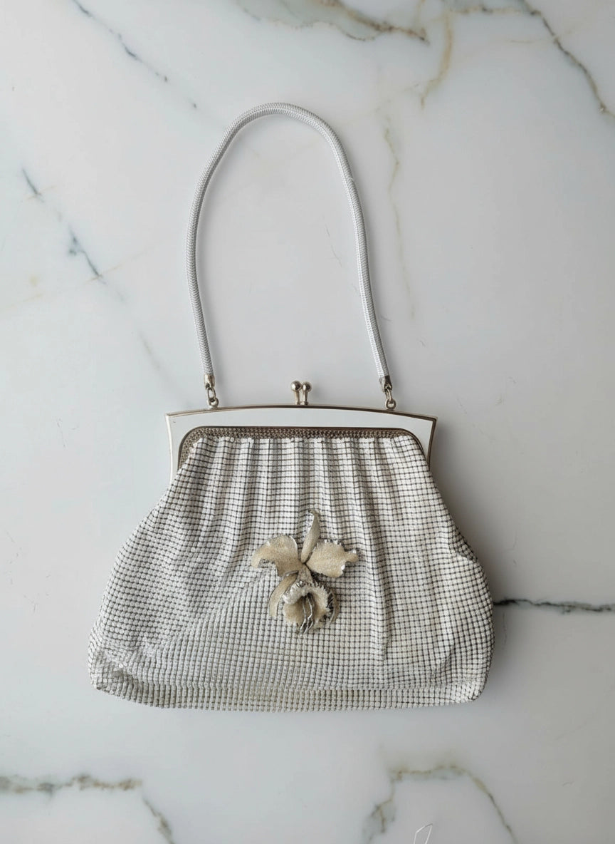 A Broochella Maison Broche l'orchidee vintage white glomesh clutch purse with a white floral brooch and white shoulder straps. The background is white marble