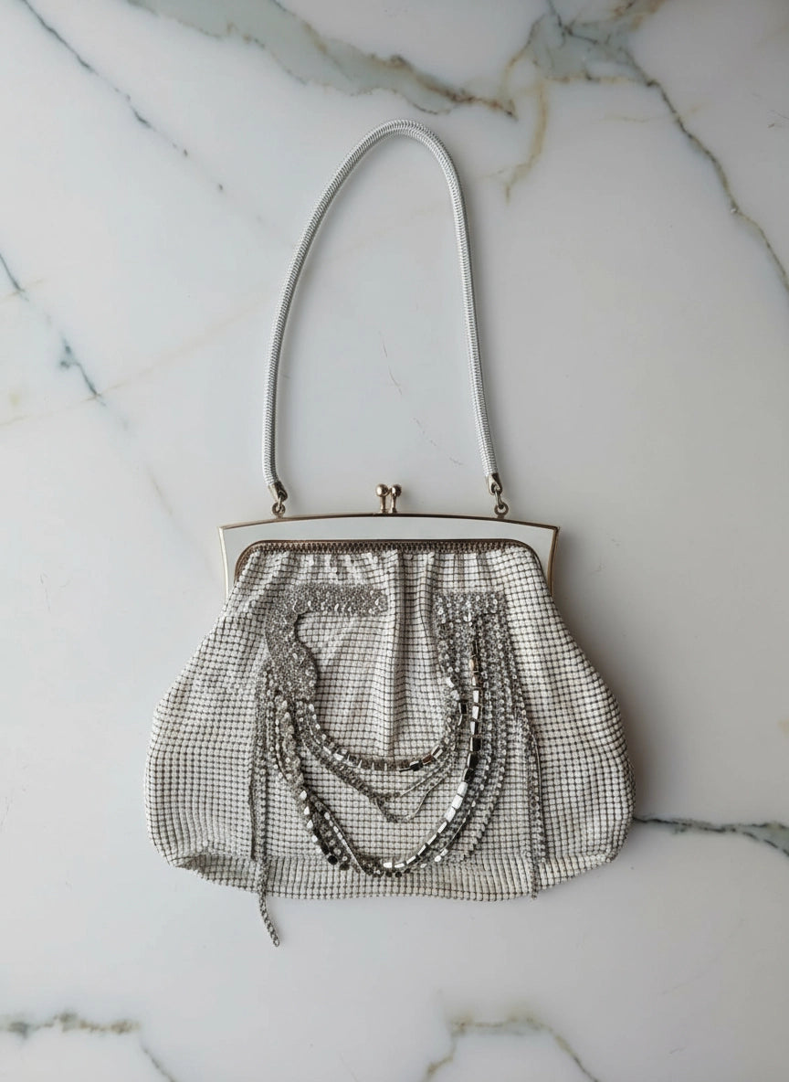 A Broochella Maison Broche l'orchidee vintage white glomesh clutch purse with a white floral brooch and white shoulder straps. The background is white marble