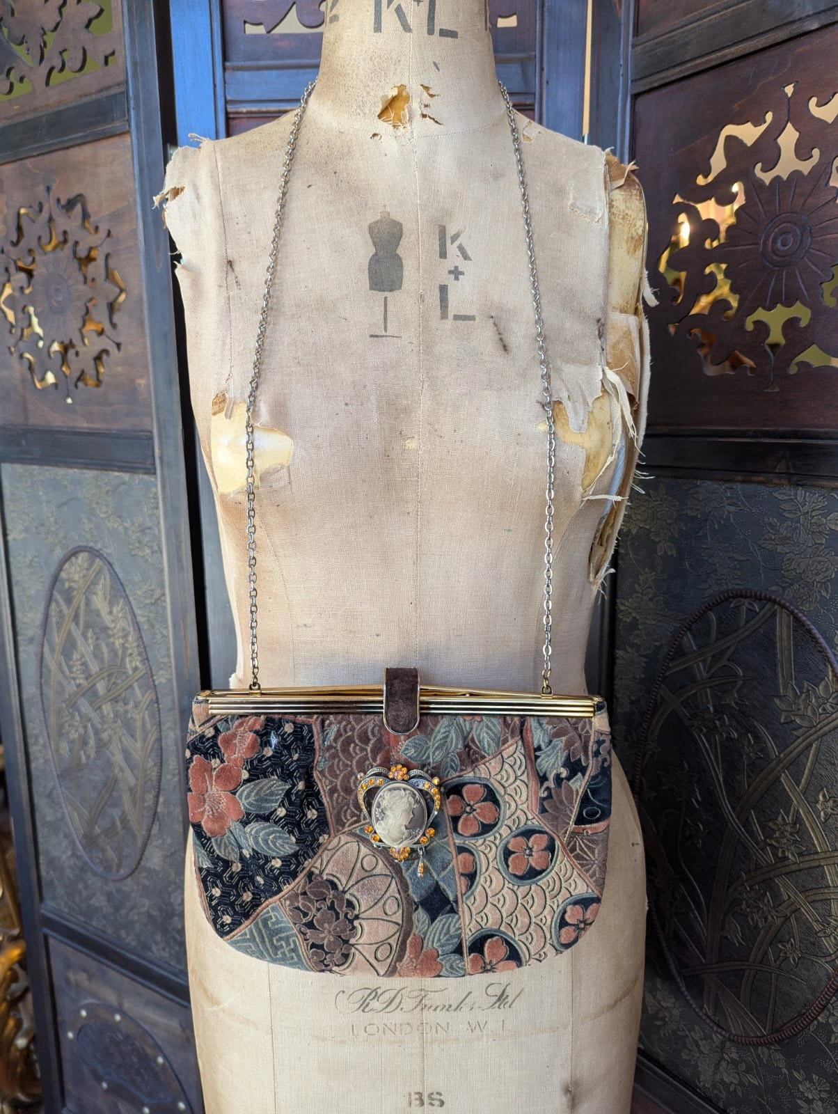 Broochella Maison Broche la cameo du jardin vintage, velvet, patterned handbag in shades of beige, blue and pink. There is a vintage cameo pinned to the bag. The bag is hanging from a vintage mannequin. The background is a decorative, brown, wooden screen. 