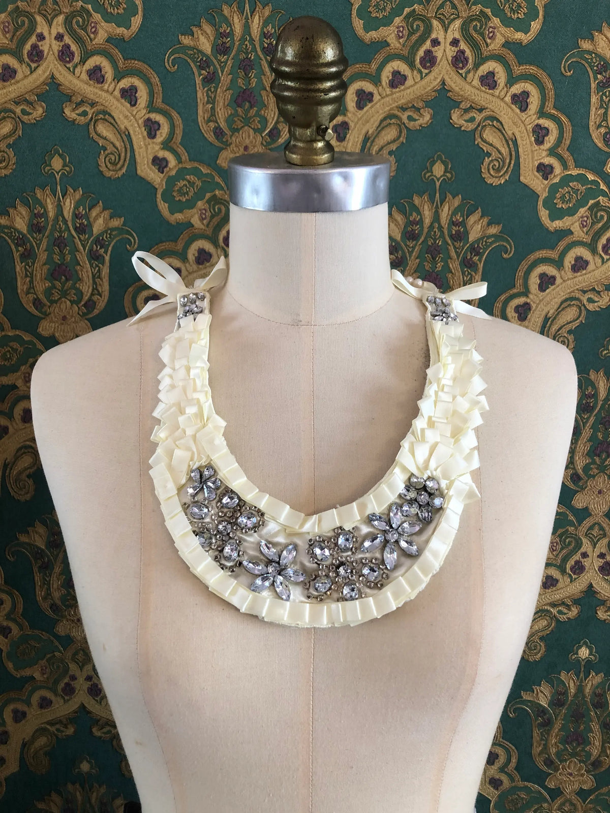 Broochella Maison Broche La Lumière Blanche ivory necklace bib with pearl strands, ivory ribbon ruffles and crystal embellishments, styled on a cream mannequin against ornate green and gold patterned wallpaper.