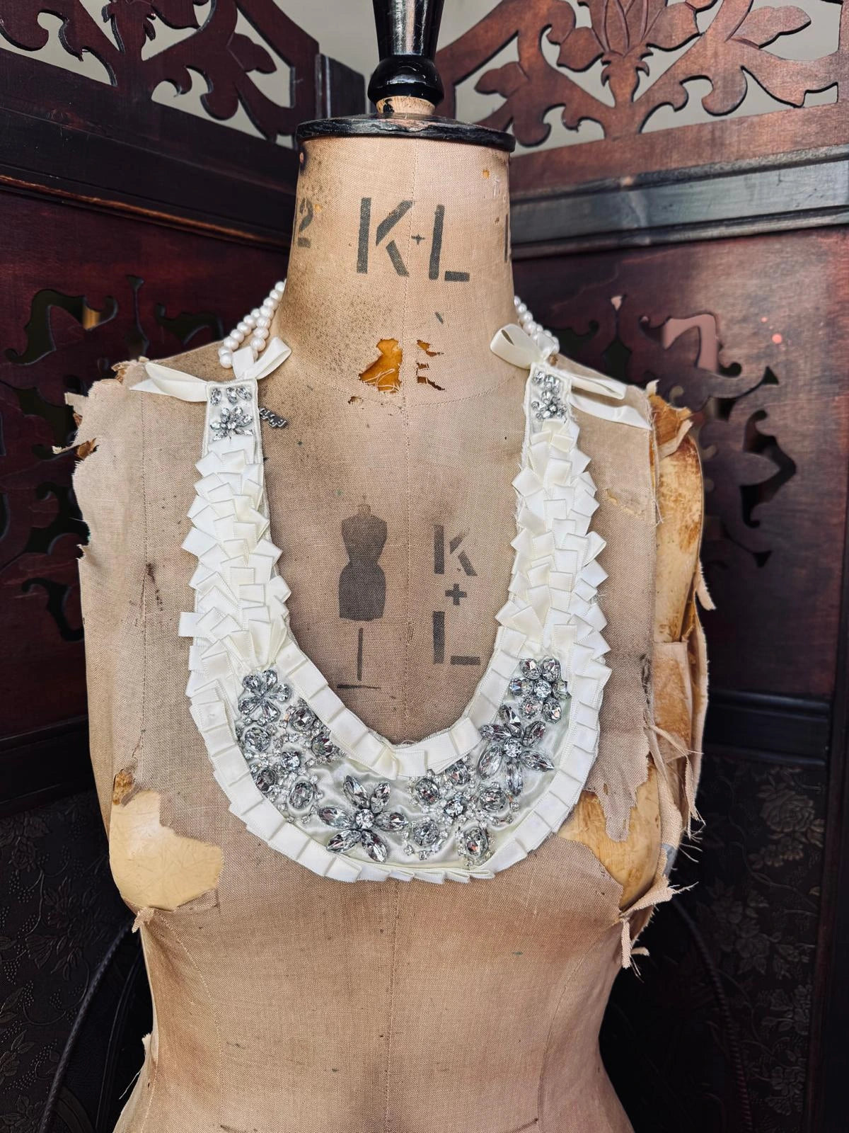 Broochella Maison Broche la Lumiere blanche handamde, ivory satin, crystal, embellished bib/necklace. Pictured with the Broochella freshwater pearl necklace. It is presented on a vintage mannequin. The background is a decorative, wooden screen. 