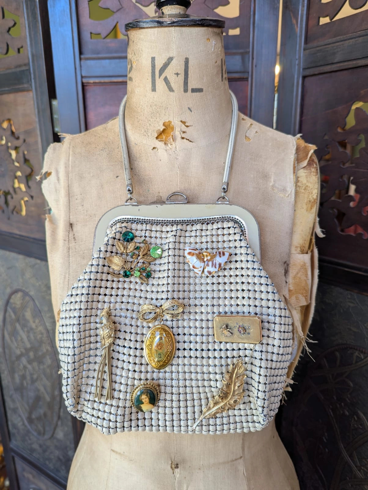 A vintage Broochella Maison Broche la memoire doree glomesh handbag with a beige mesh bead body, a silver-tone metal frame, and a matching shoulder strap. It is embellished with several gold toned vintage brooches.  It is displayed on a vintage mannequin. The background is a decorative, brown, wooden panel. 