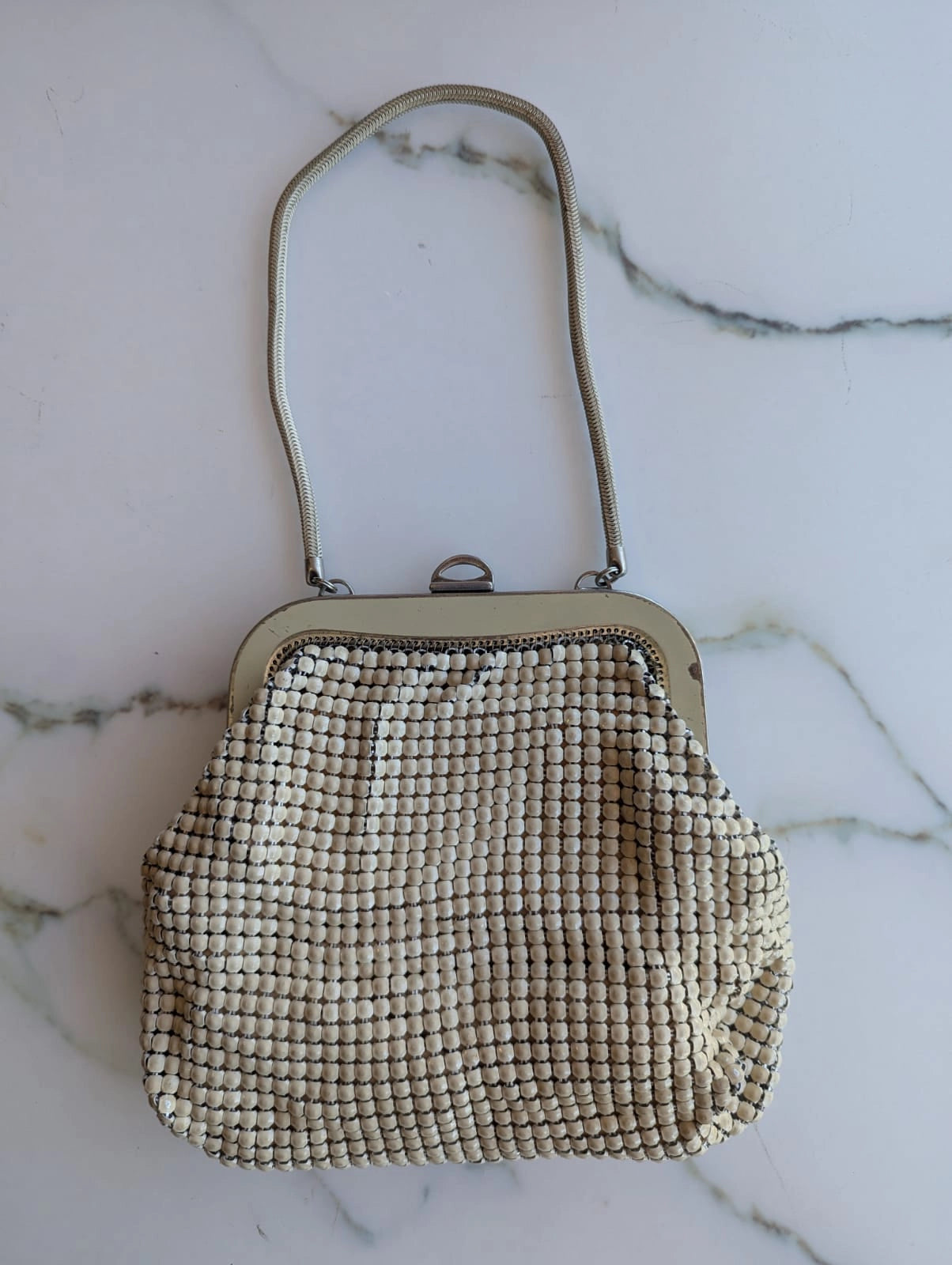 A vintage Broochella Maison Broche la memoire doree glomesh handbag with a beige mesh bead body, a silver-tone metal frame, and a matching shoulder strap, displayed on a white marble surface.