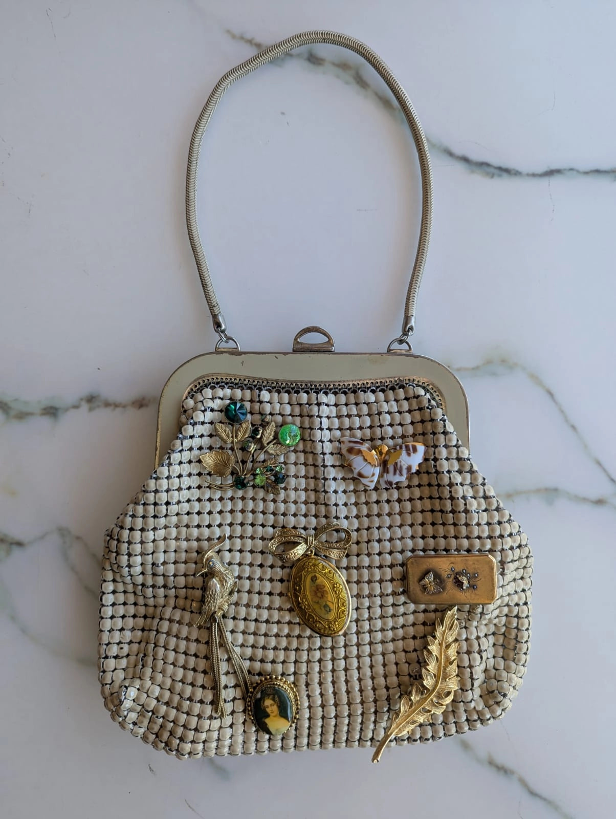 A vintage Broochella Maison Broche la memoire doree glomesh handbag with a beige mesh bead body, a silver-tone metal frame, and a matching shoulder strap. It is embellished with several gold toned vintage brooches.  It is displayed on a white marble surface.