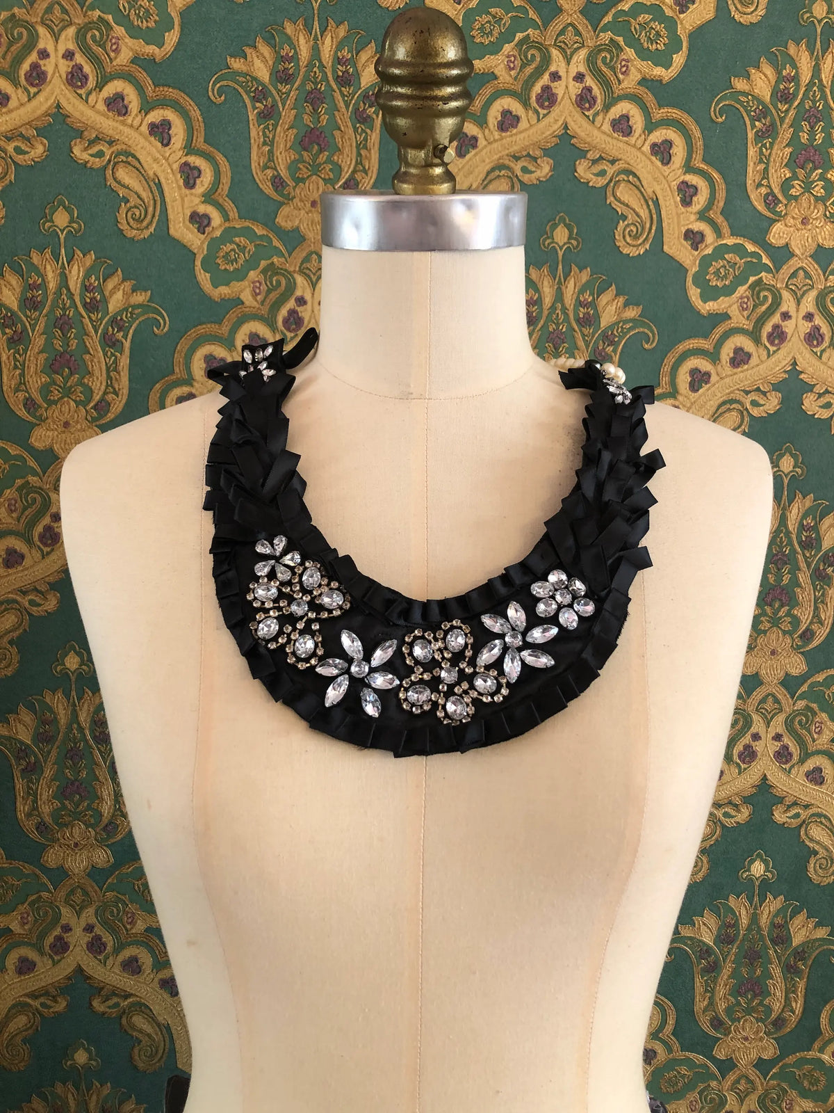 Broochella Maison Broche L’Amour Paré black statement necklace bib styled on a cream dressmaker’s mannequin, featuring layered black ribbon, sparkling clear crystal floral embellishments and ivory pearl strands, against an ornate dark green and gold patterned textile background.