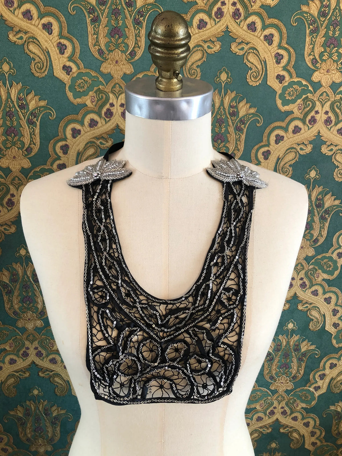 Broochella Maison Broche Le Coeur black necklace bib with intricate black lace, silver sequin detailing and embellished silver leaf motifs, styled on a cream mannequin against ornate green and gold patterned wallpaper.