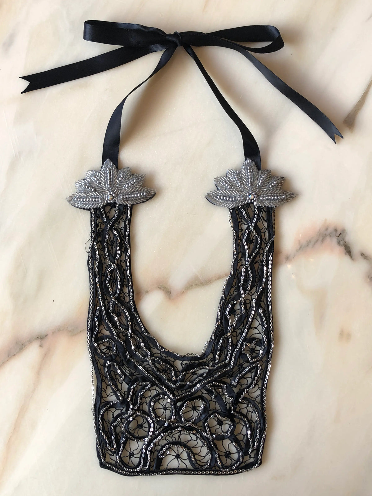 Broochella Maison Broche Le Coeur black necklace bib featuring intricate lace, silver sequin embroidery, embellished silver leaf motifs and a black satin ribbon tie, photographed on a pale white marble background.
