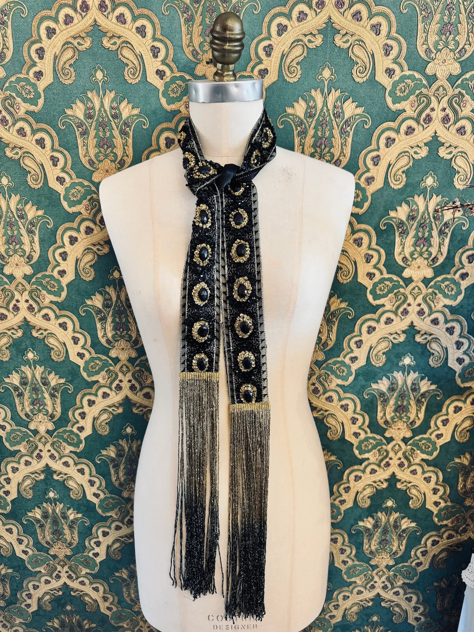 Broochella Antionette handmade fringe black and gold embellished foulard neck tie. Pictured on a mannequin. The background is green and gold damask wallpaper.
