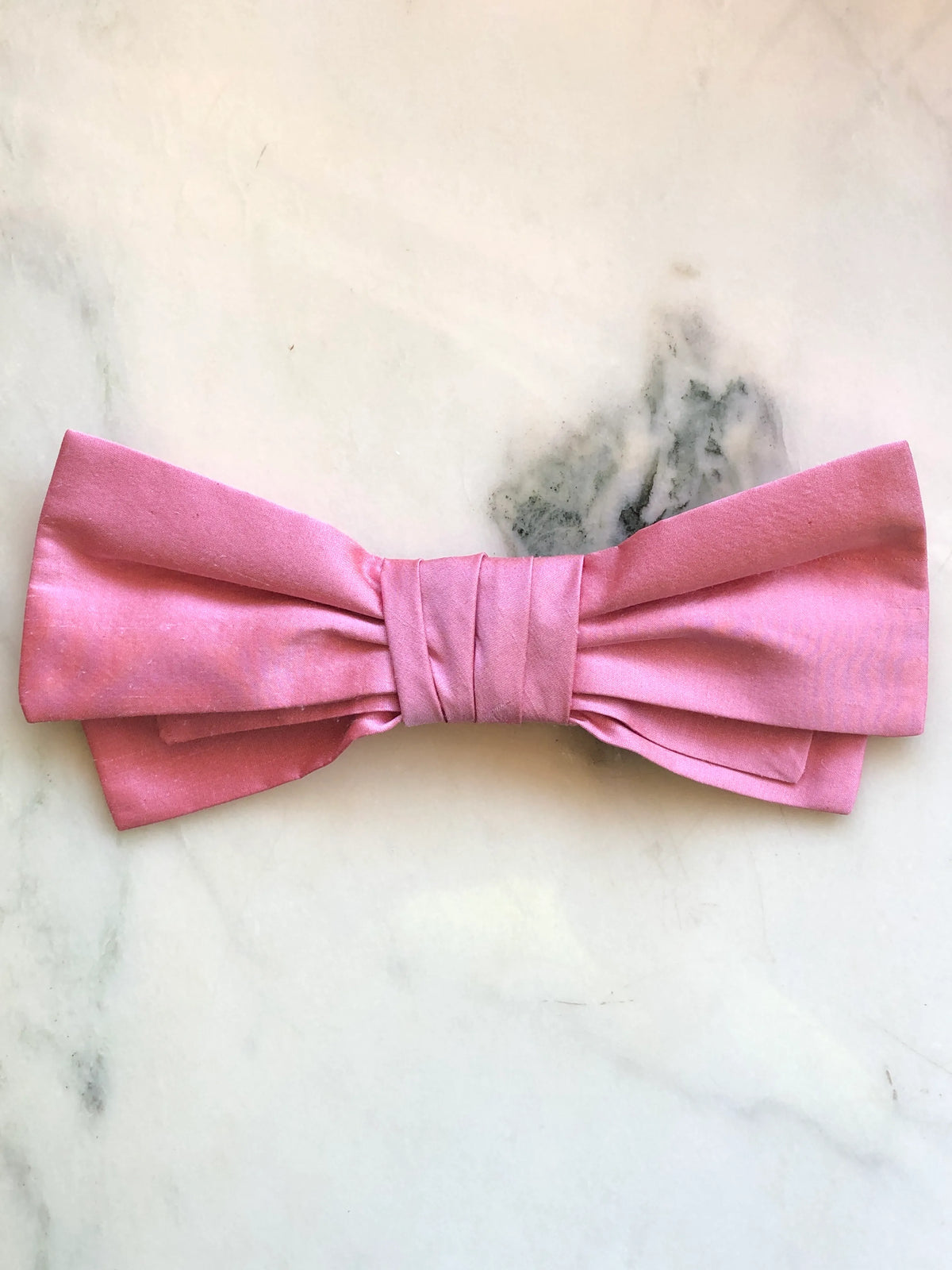 Broochella Marie Antoinette pink silk bow brooch pin in soft rose pink with a structured double bow and pleated wrapped centre, photographed on a white marble background with soft grey-green veining.