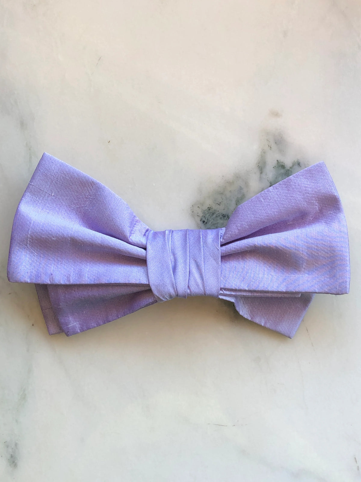 Broochella Marie Antoinette purple silk bow brooch pin with a softly structured double bow and wrapped centre, photographed on a white marble background with soft grey-green veining.