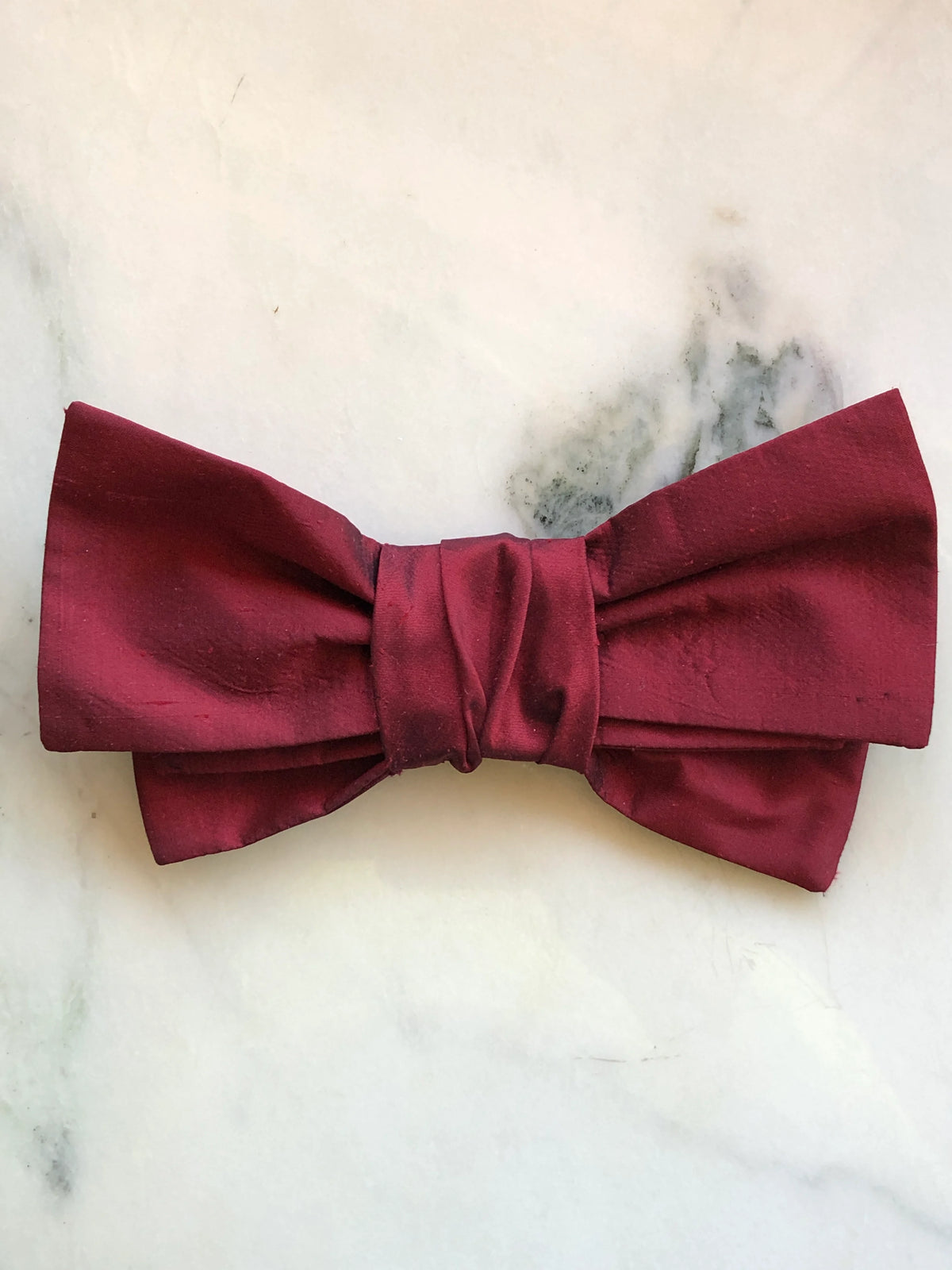 Broochella Marie Antoinette red silk bow brooch pin in deep crimson red with subtle diamond-textured silk, a double bow silhouette and wrapped centre, photographed on a white marble background with soft grey-green veining.