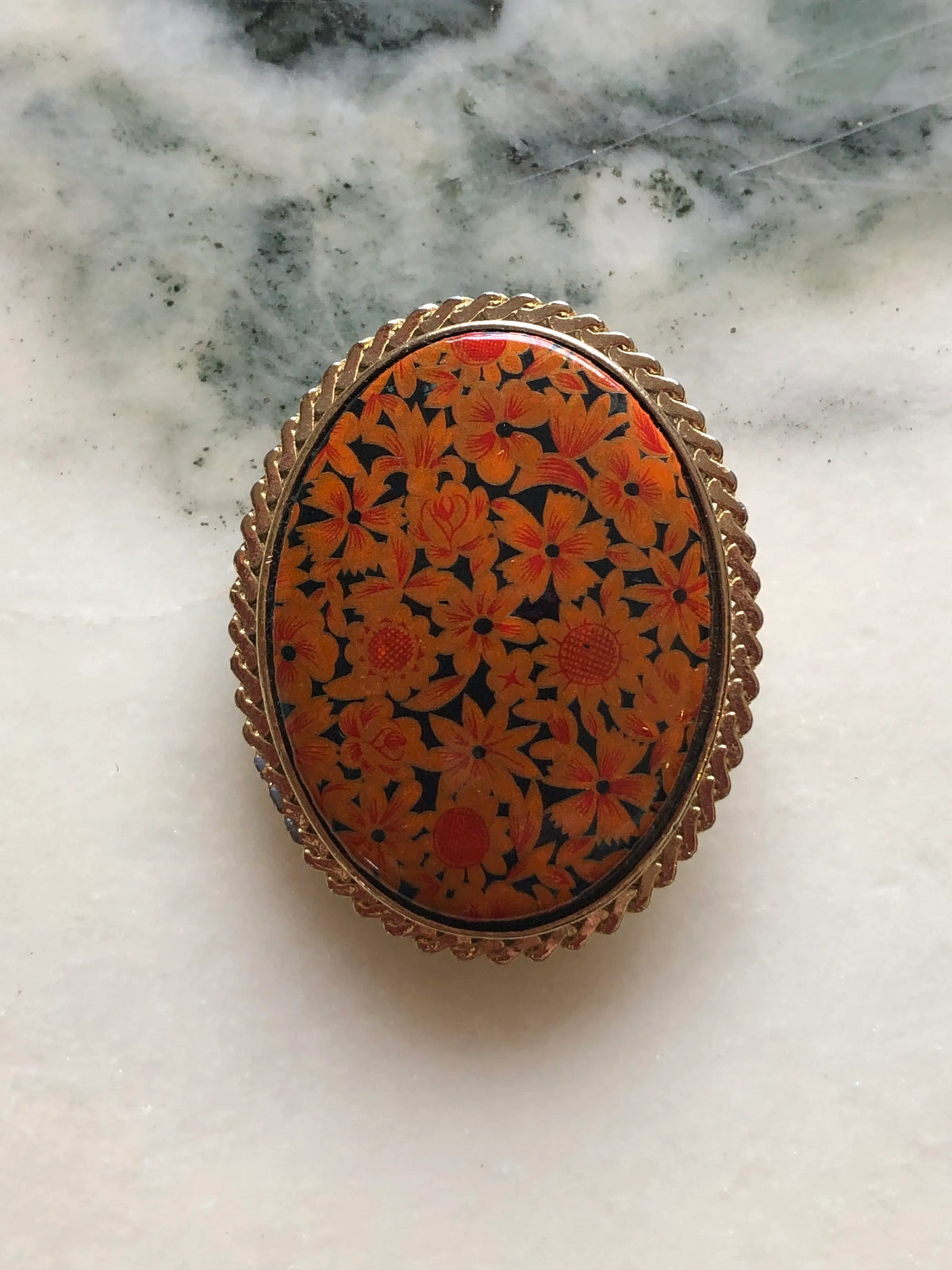 Broochella Marigold Garden vintage oval brooch with orange and amber flowers on a black background, a glossy domed centre and textured gold-tone frame, measuring approximately 4.5 × 3.5 cm. Pictured on white and grey marble