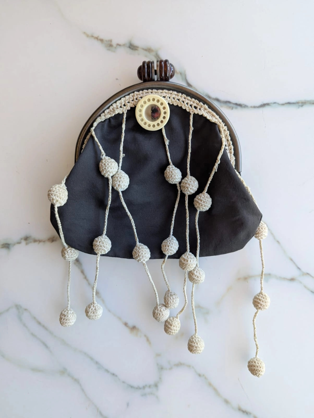 A Broochella Marni grey cotton kiss-lock clutch purse with cream-coloured crochet fringe beads hanging from the clasp, displayed on a white marble surface.