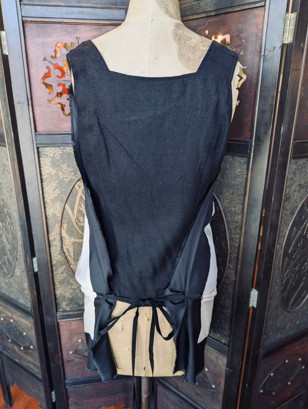The back view of a Max Mara Bib Top Silk  – Size 38. White and black draped bib front top with a back tie. It is pictured on a mannequin. The background is an ornate, dark wood room divider