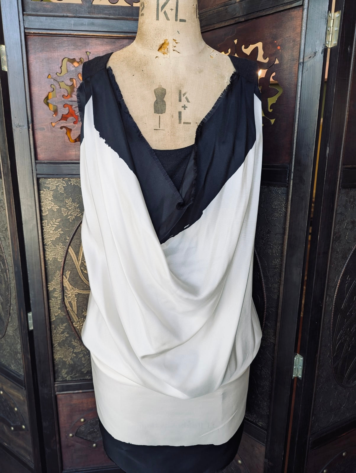 Max Mara Bib Top Silk  – Size 38. White and black draped bib front top with a back tie. It is pictured on a mannequin. The background is an ornate, dark wood room divider