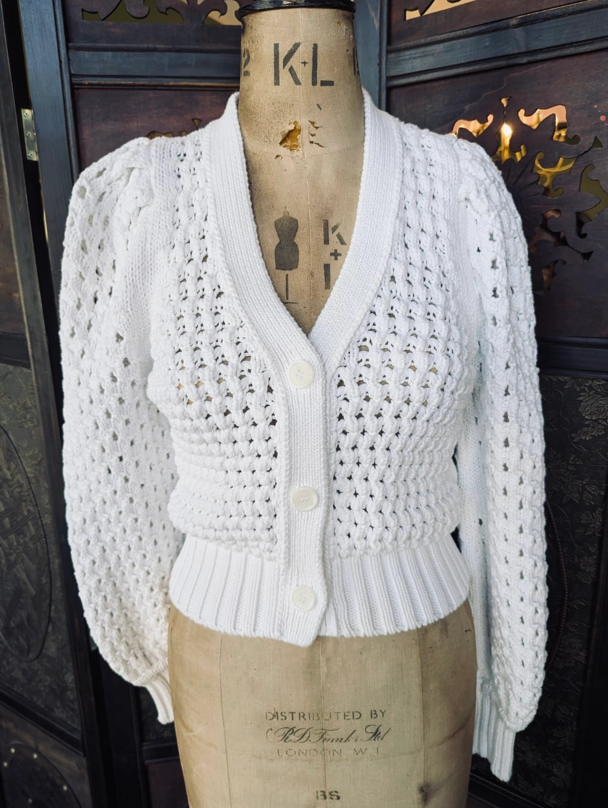 Broochella Me + Em cotton crochet knit cardigan in white. It is pictured on a vintage mannequin. The background is an ornate dark wood room divider.