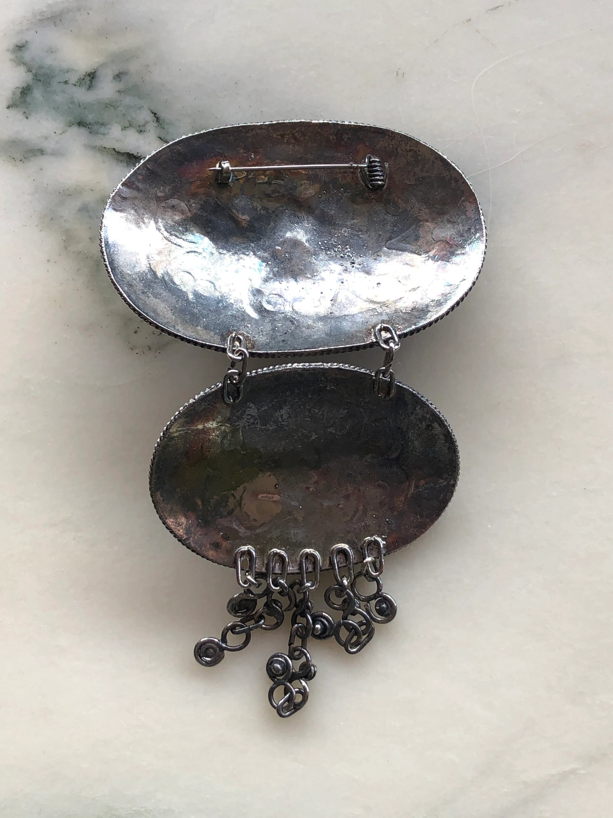 Broochella Medaglione Cascata Vintage Large Brooch Pin back view showing the two connected oval medallions, traditional pin fastening and dangling metal charms, photographed on a white marble background with soft grey-green veining.