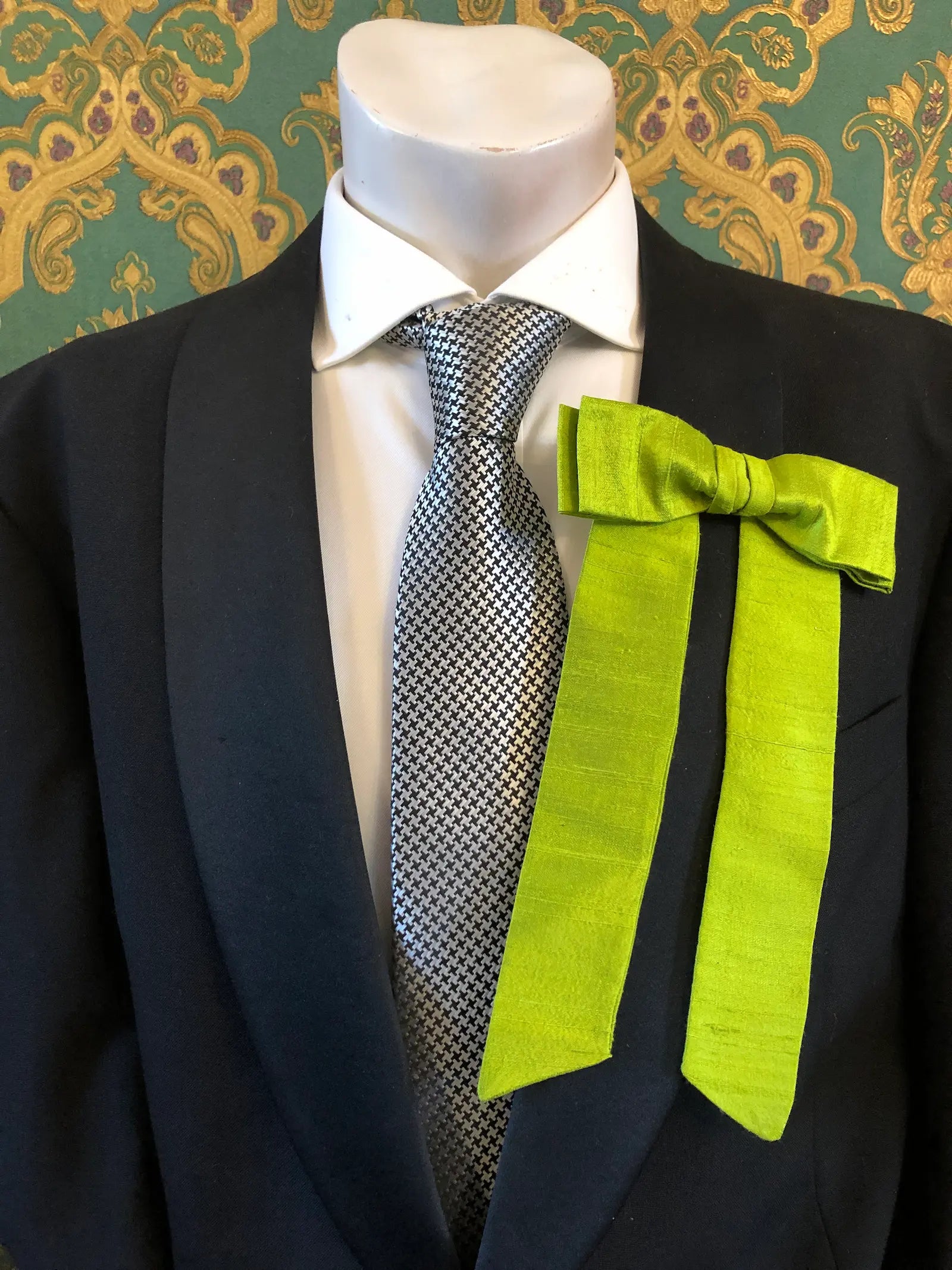 A Broochella Men's Alice silk bow brooch in lime green. It is styled on a white shirt and patterned tie, displayed on a black suit jacket mannequin torso against a green and gold damask wallpaper background.