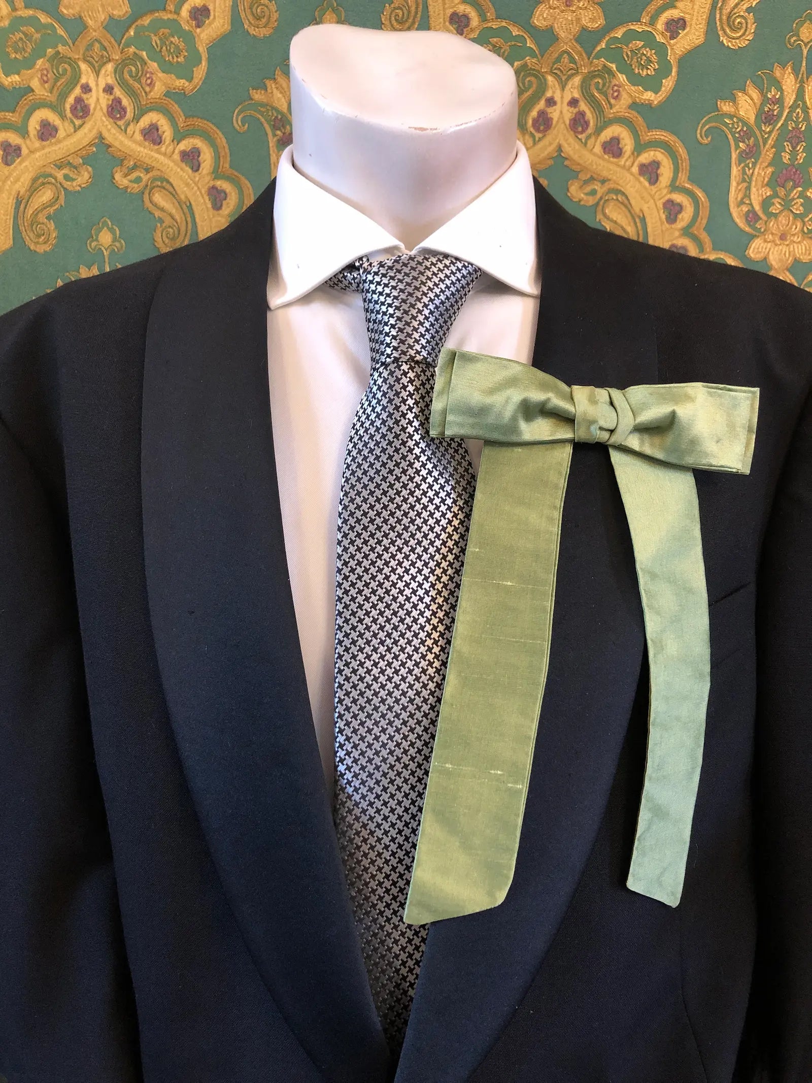 A Broochella Men's Alice silk bow brooch in avocado. It is styled on a white shirt and patterned tie, displayed on a black suit jacket mannequin torso against a green and gold damask wallpaper background.