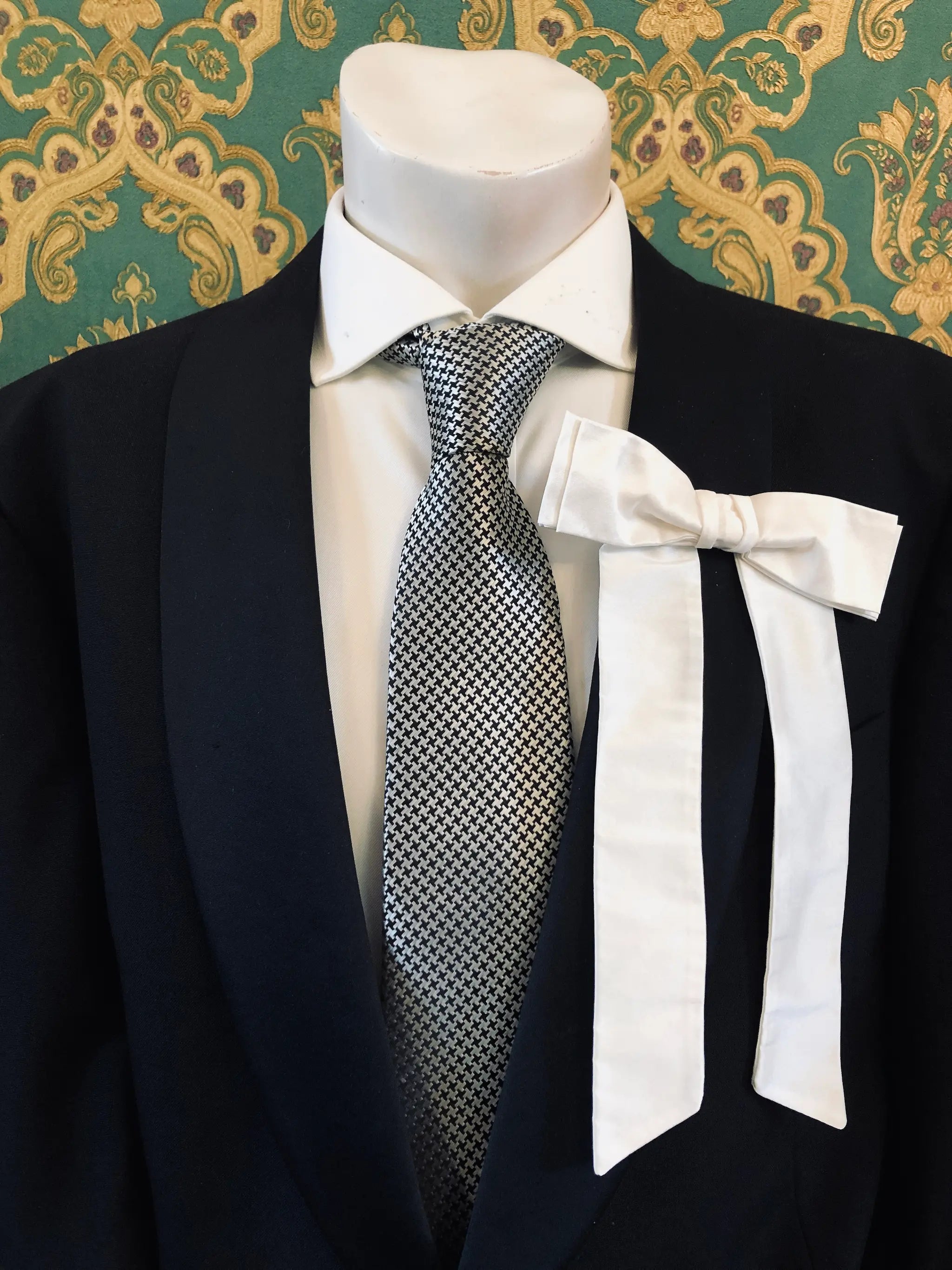 A Broochella Men's Alice silk bow brooch in ivory. It is styled on a white shirt and patterned tie, displayed on a black suit jacket mannequin torso against a green and gold damask wallpaper background.