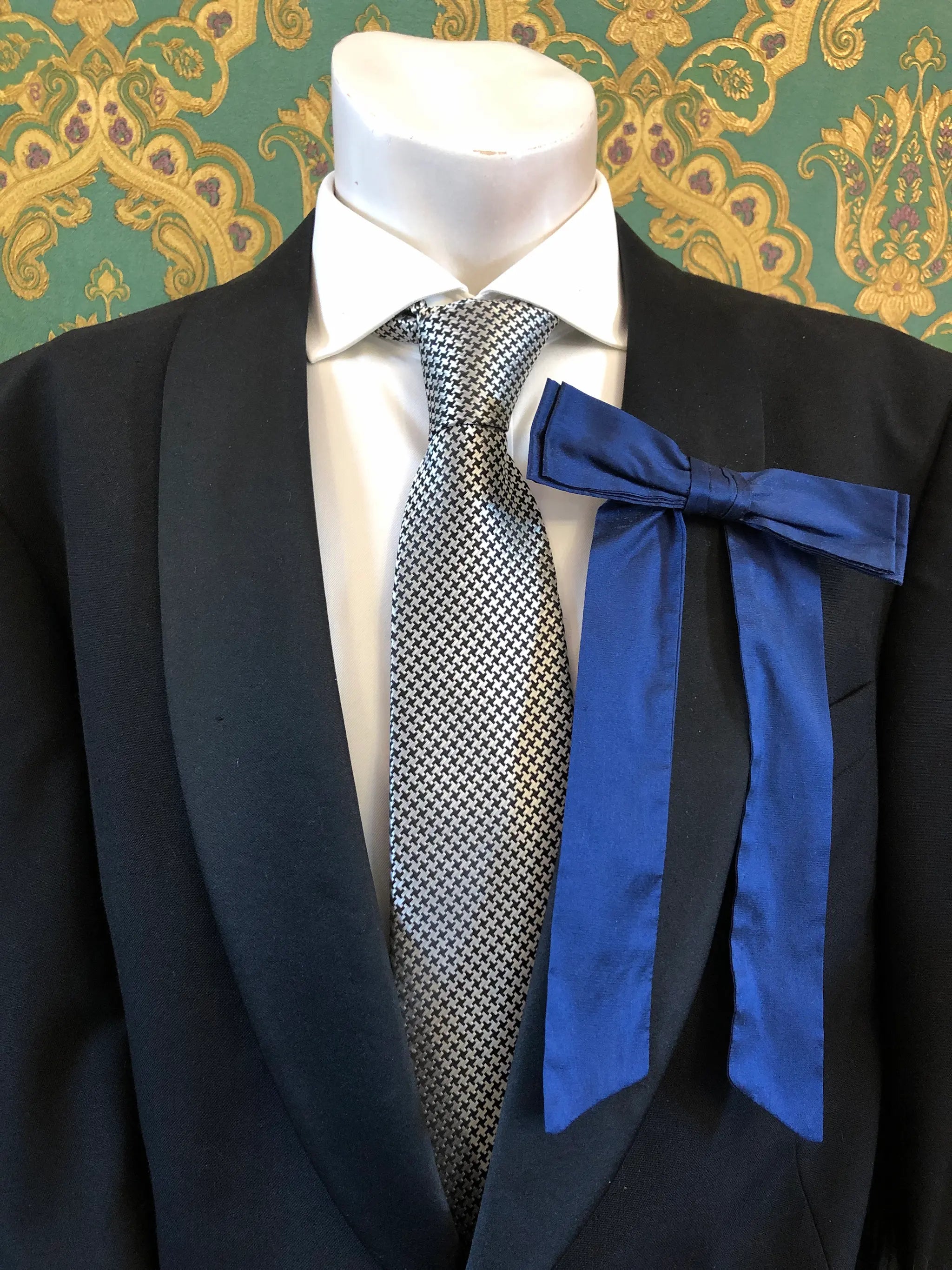 A Broochella Men's Alice silk bow brooch in navy blue. It is styled on a white shirt and patterned tie, displayed on a black suit jacket mannequin torso against a green and gold damask wallpaper background.