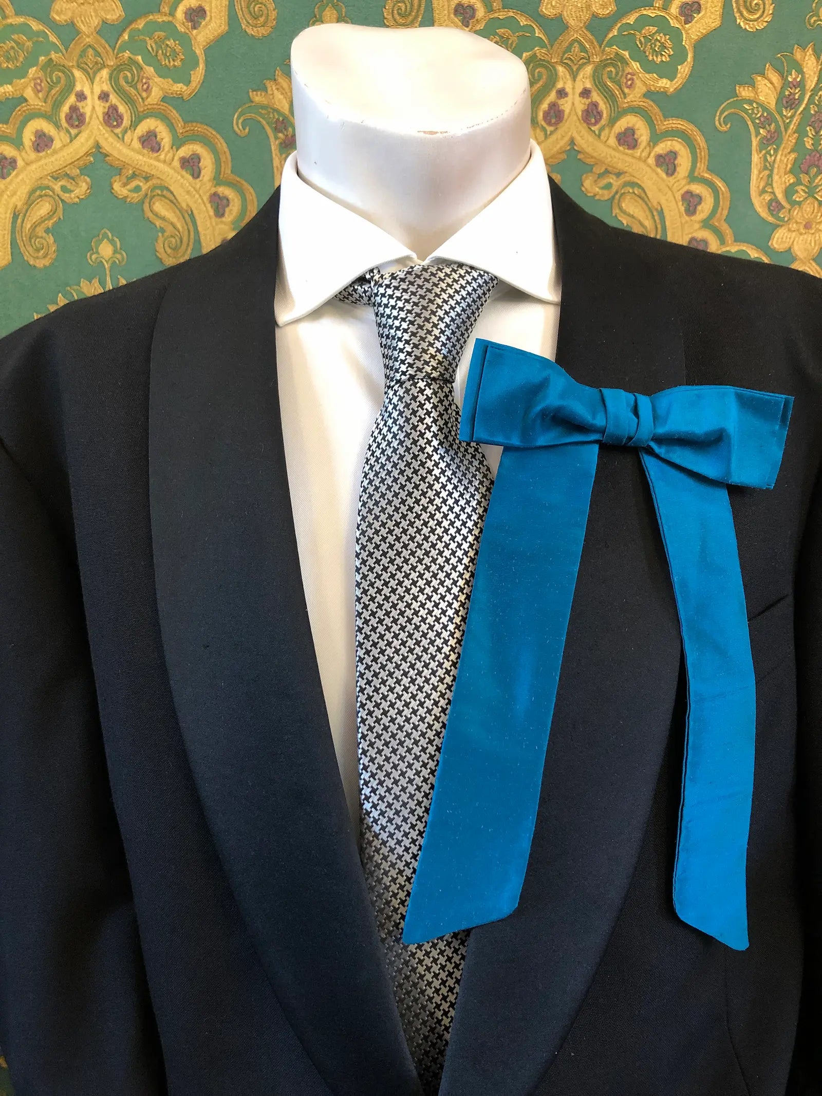 A Broochella Men's Alice silk bow brooch in turquoise. It is styled on a white shirt and patterned tie, displayed on a black suit jacket mannequin torso against a green and gold damask wallpaper background.