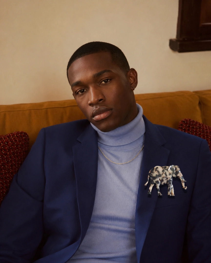 A man wearing a navy blue blazer over a light blue turtleneck sweater, accessorised with a gold chain necklace and a Broochella Men's Animalia tortoiseshell elephant-shaped lapel pin, seated on a mustard yellow couch