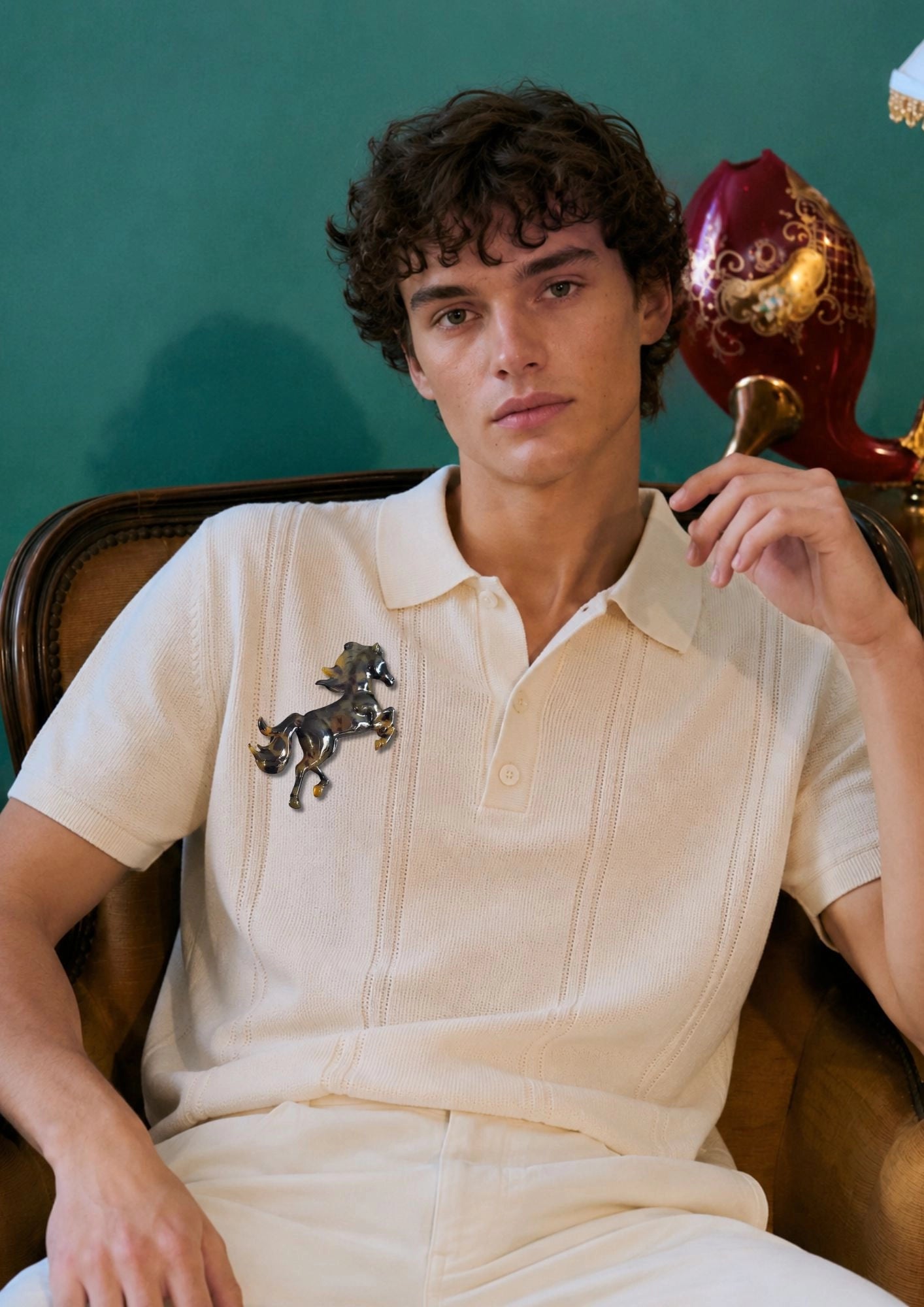 A man wearing a cream-colored short-sleeve polo shirt with a Broochella Men's Animalia tortoiseshell horse brooch on the chest, seated in a brown leather armchair against a dark green wall.