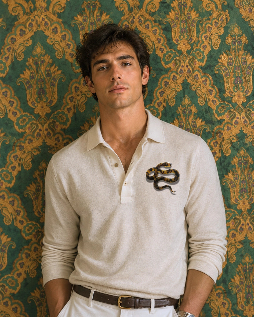 A dark haired male model leans against a green and gold damask wallpaper background. He is wearing a beige long sleeve polo shirt, a brown belt and beige chinos. Pinned to his chest is the Broochella Men's Animalia tortoiseshell snake brooch in brown. 