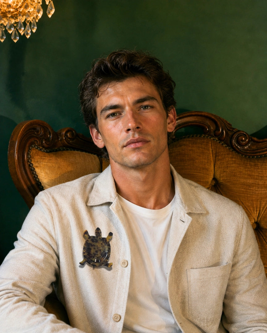 A man wearing a cream-coloured linen jacket over a white t-shirt, seated in an ornate wooden armchair against a dark green wall. Pinned to his shirt is the Broochella Men's Animalia Tortoiseshell Tortoise Brooch in Brown. 