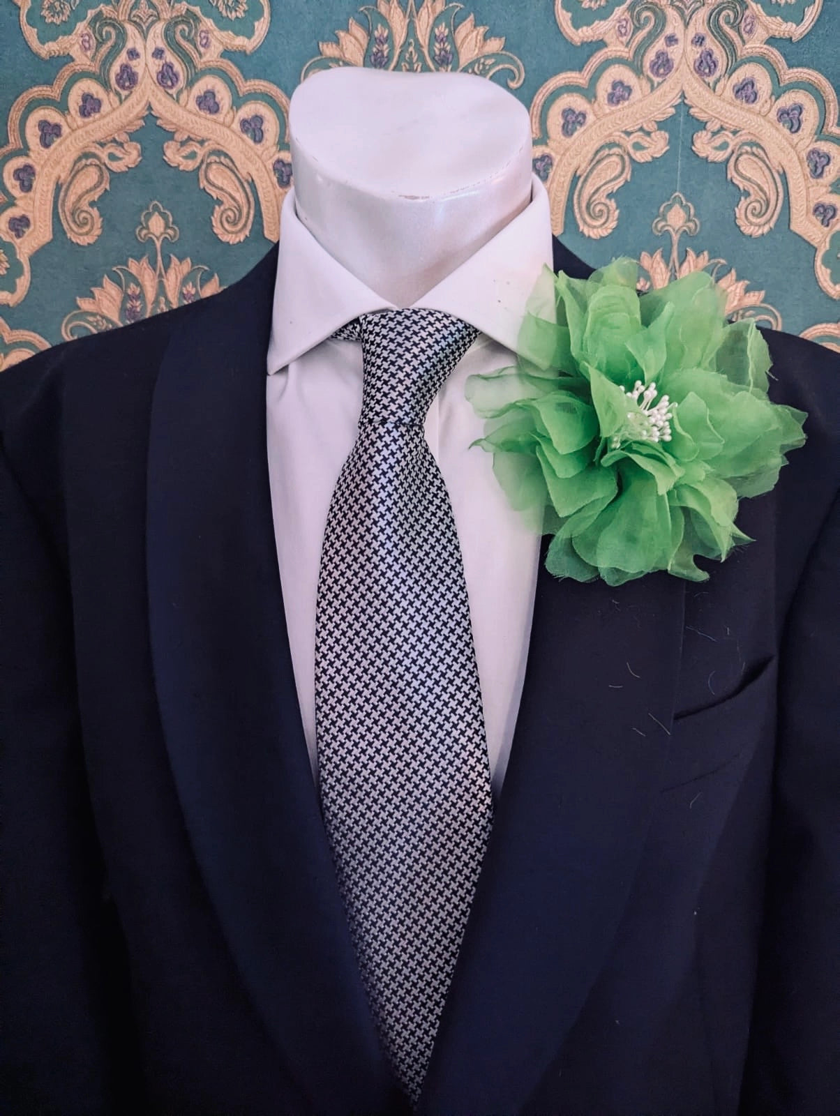 A black suit jacket displayed on a white mannequin torso, styled with a white dress shirt, a patterned grey and white tie, and a Broochella men's green Aubrieta flower brooch against a teal and gold damask wallpaper background.