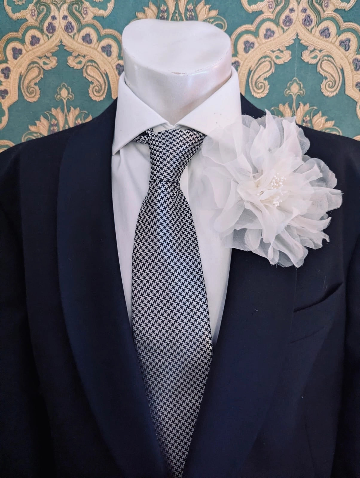 A black suit jacket displayed on a white mannequin torso, styled with a white dress shirt, a patterned grey and white tie, and a Broochella men's ivory Aubrieta flower brooch against a teal and gold damask wallpaper background.
