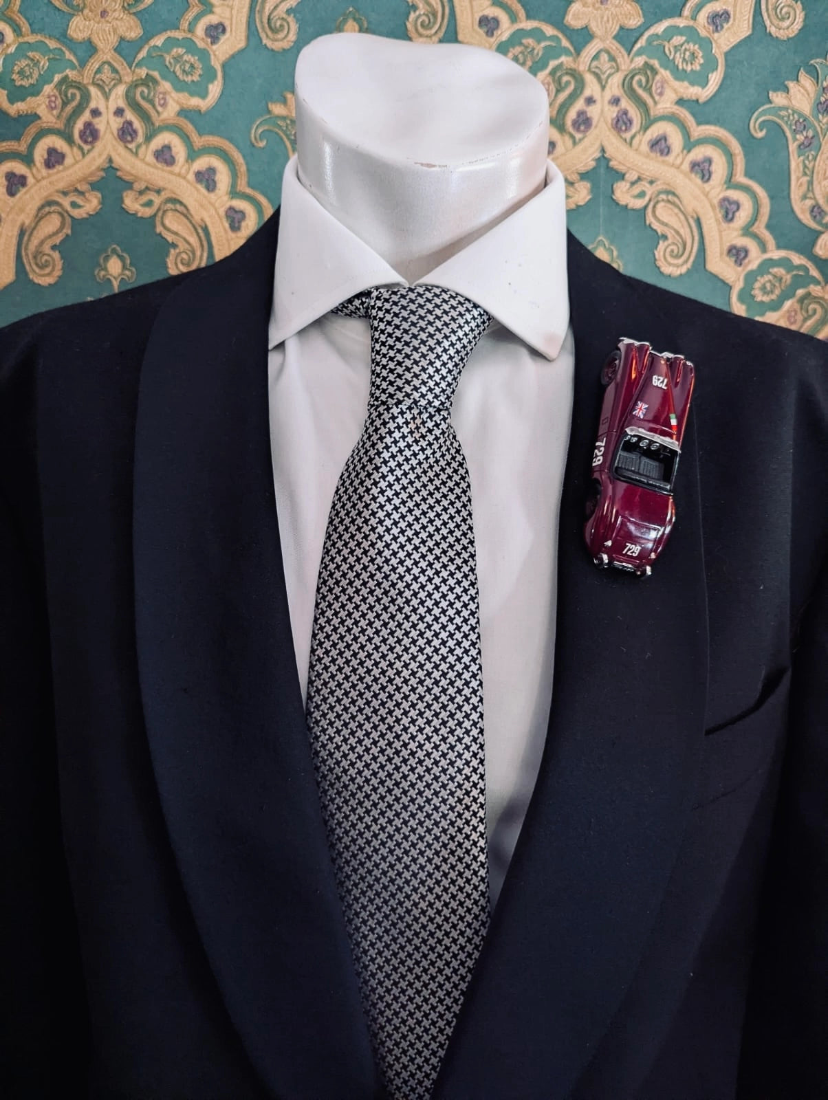 Broochella men's scuderia d'oro barolo 729 burgundy vintage formula one racing car brooch. Pinned on a men's mannequin wearing a black suit and a houndstooth tie. The background is green and gold damask wallpaper. 