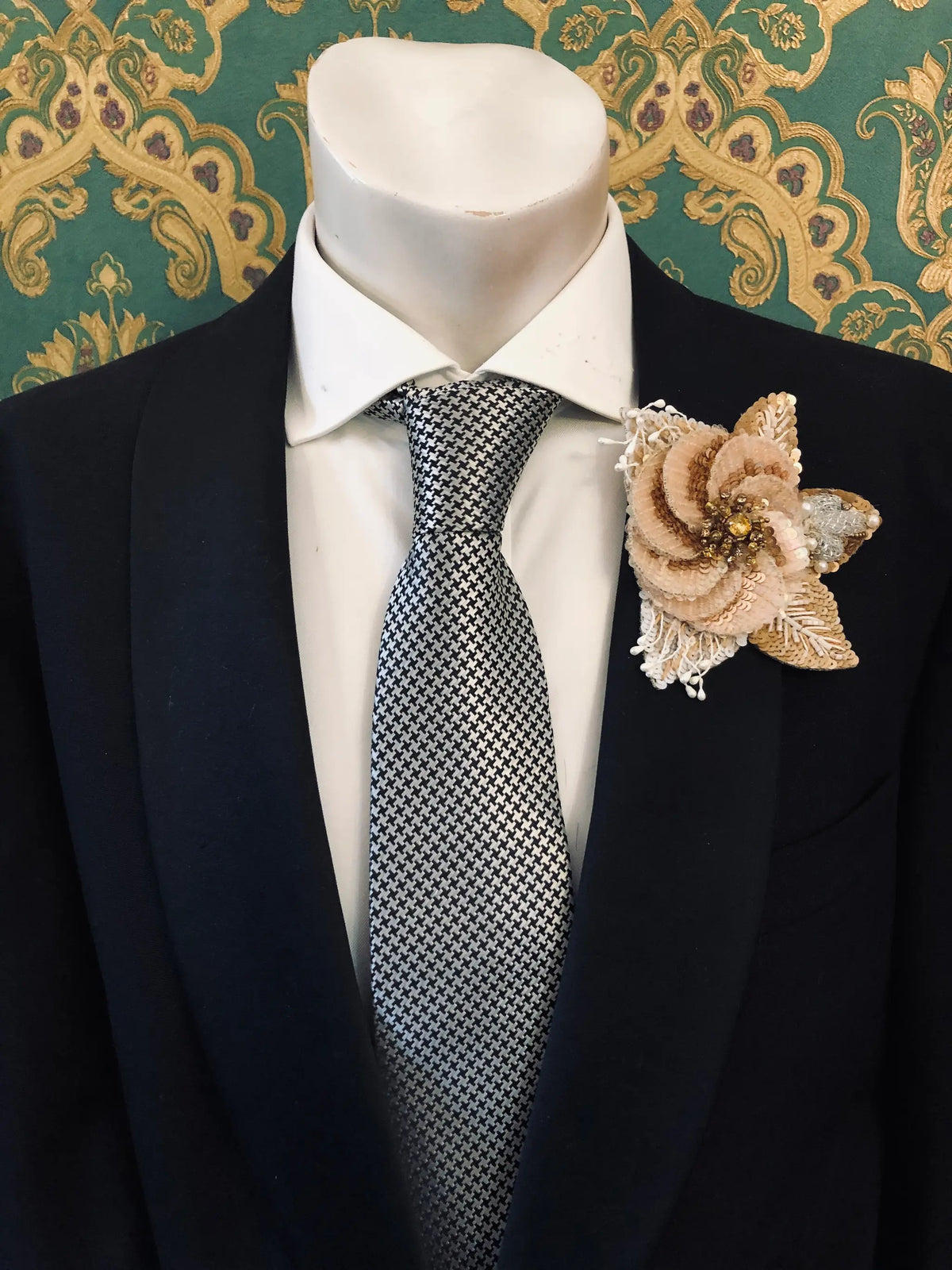 A men's formal outfit displayed on a white mannequin torso, featuring a black suit jacket, a white dress shirt, a silver and black checkered tie, and a Broochella Blossom de Luxe beige sequinned fabric flower brooch