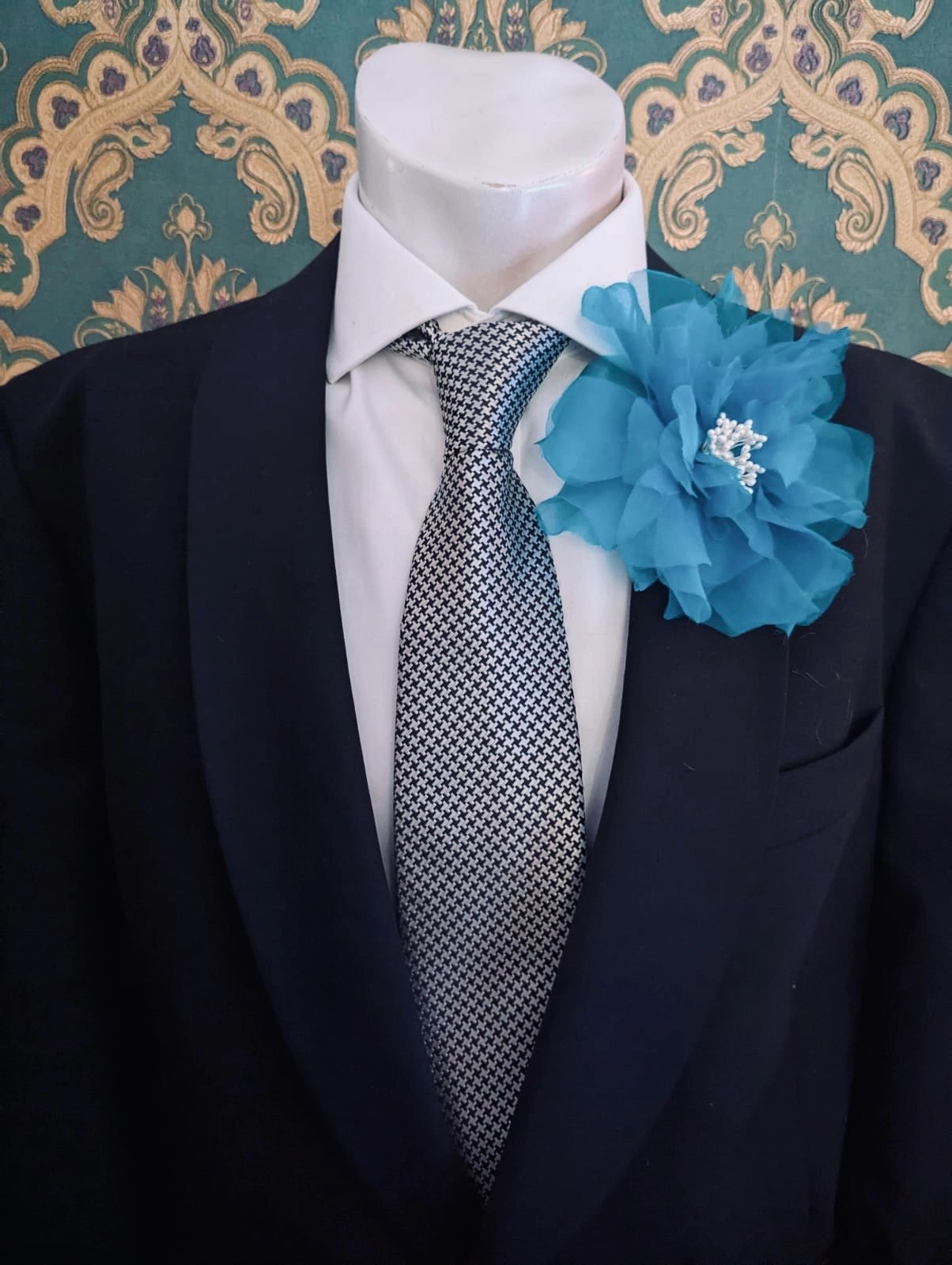 A black suit jacket displayed on a white mannequin torso, styled with a white dress shirt, a patterned grey and white tie, and a Broochella men's blue Aubrieta flower brooch against a teal and gold damask wallpaper background.