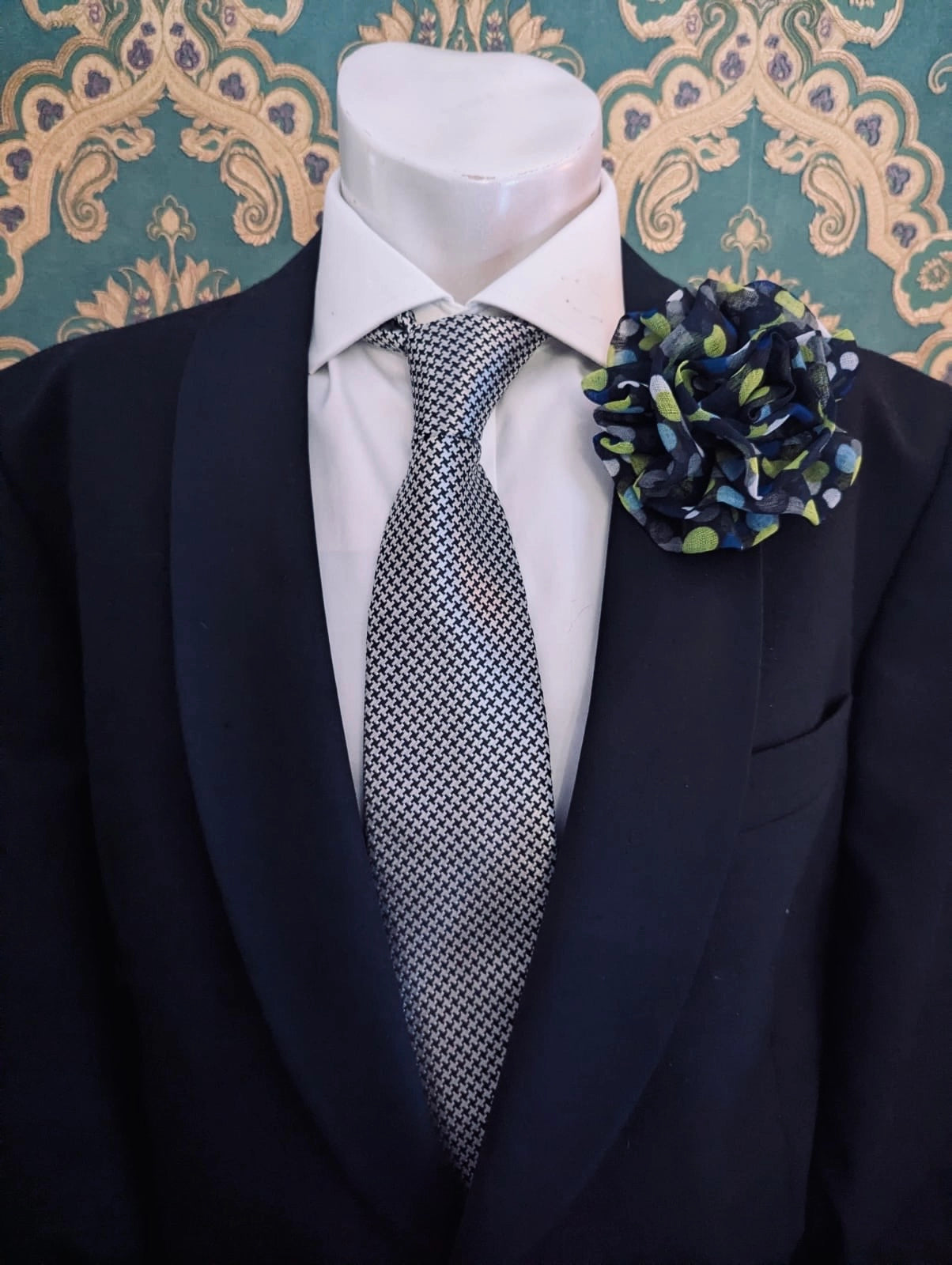 A men's formal outfit displayed on a white mannequin torso, featuring a black suit jacket, a white dress shirt, a grey and white patterned tie, and the Broochella brassica men's flower brooch in navy and green polka dots on the lapel, set against a teal and gold damask wallpaper background.