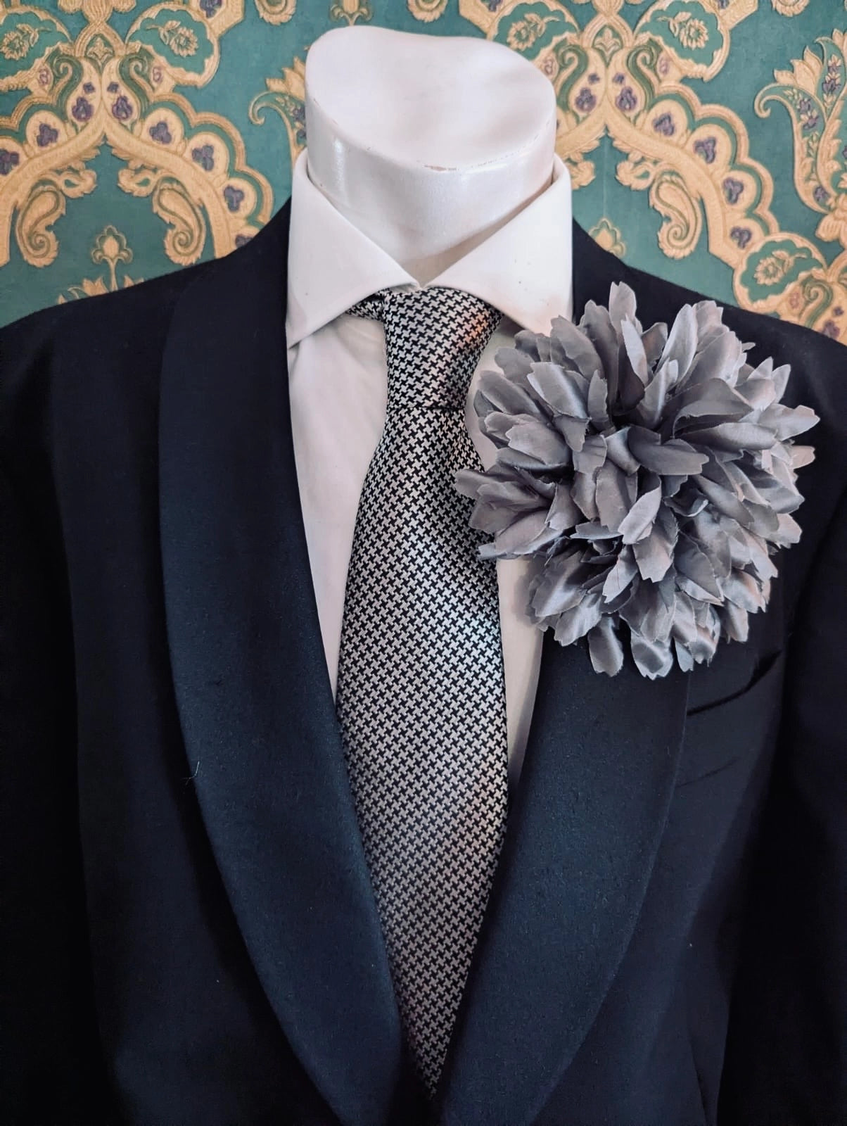 Mannequin wearing a black suit with a white shirt, patterned tie and Broochella calendula fabric grey flower brooch against a decorative wall.