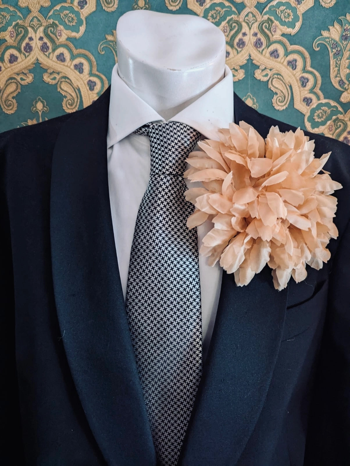Mannequin wearing a black suit with a white shirt, patterned tie and Broochella calendula fabric peach flower brooch against a decorative wall.