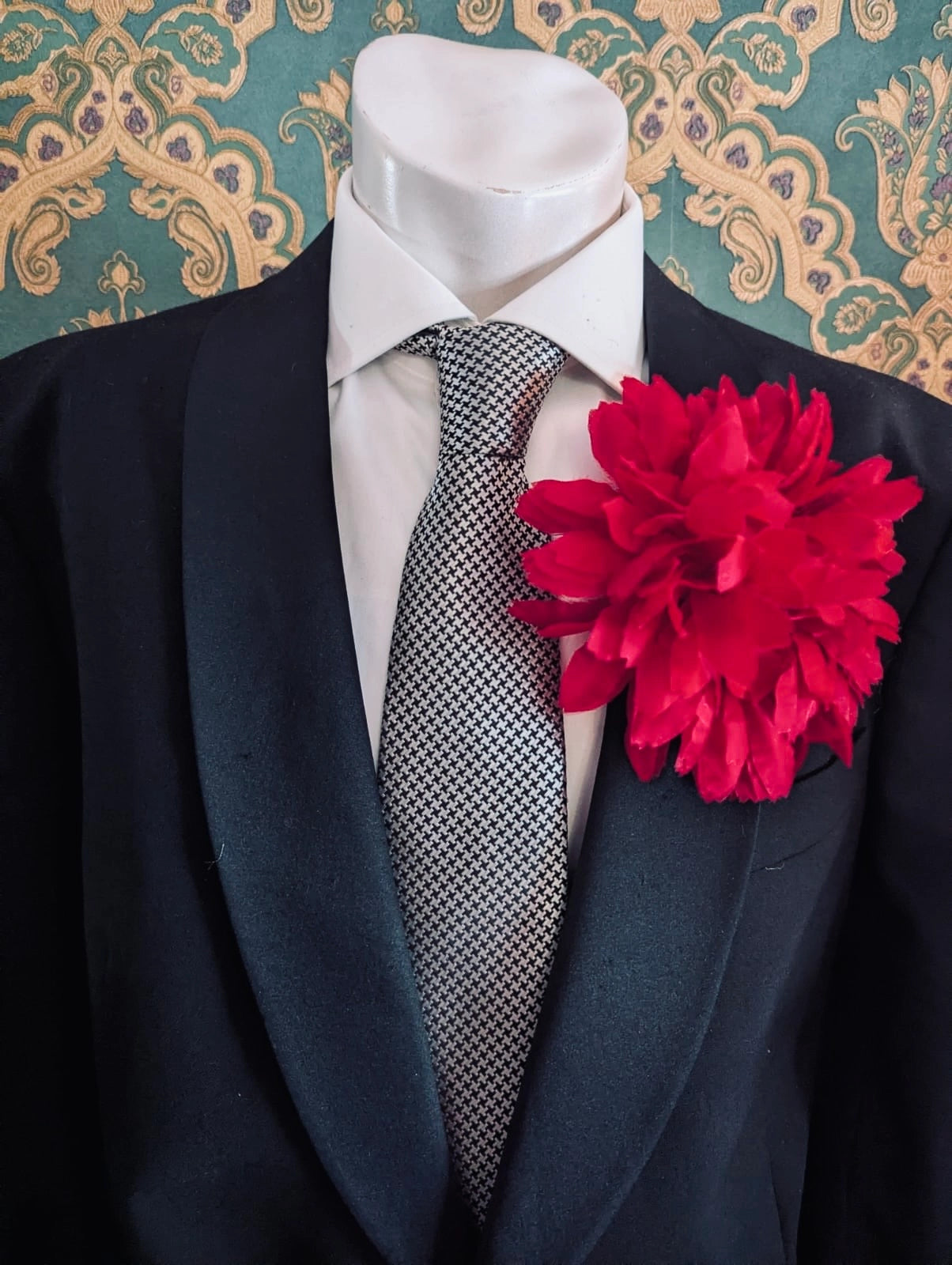 Mannequin wearing a black suit with a white shirt, patterned tie and Broochella calendula fabric red flower brooch against a decorative wall.
