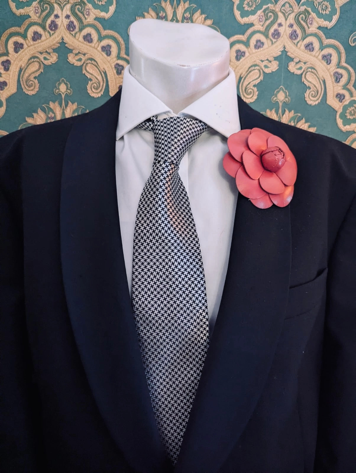 A mannequin torso wearing a black suit jacket, white dress shirt, and patterned gray tie, with a Broochella men's camelia coral-pink leather flower brooch on the left lapel. Ornate green and gold patterned wallpaper is visible in the background.