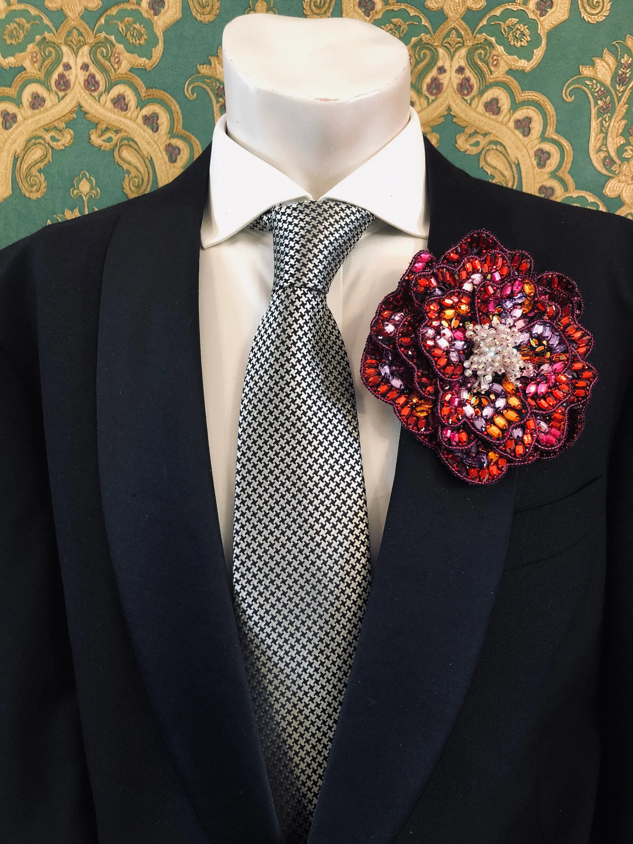 The Broochella Men's Cristallino hand beaded designer brooch made from pink red and orange beads. It is pinned to the lapel of a men's mannequin that is wearing a black jacket, a white shirt and a grey and silver houndstooth tie. The background is green and gold damask wallpaper. 
