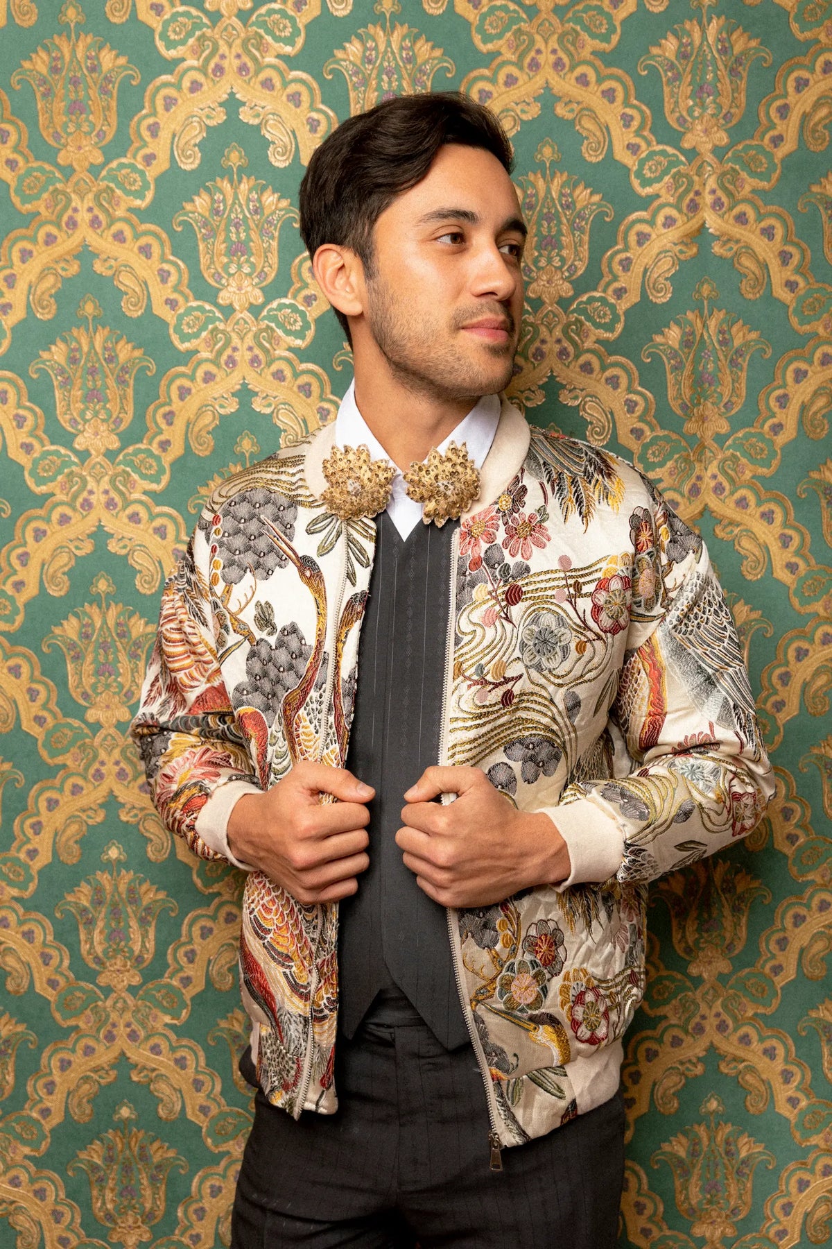 A man wearing a cream-colored bomber jacket with an intricate multicolor floral and botanical print, styled with a white shirt, black vest, and black trousers, standing against a green and gold patterned wallpaper background. He is wearing the Broochella men's foglie  d'autunno leaf brooch set.