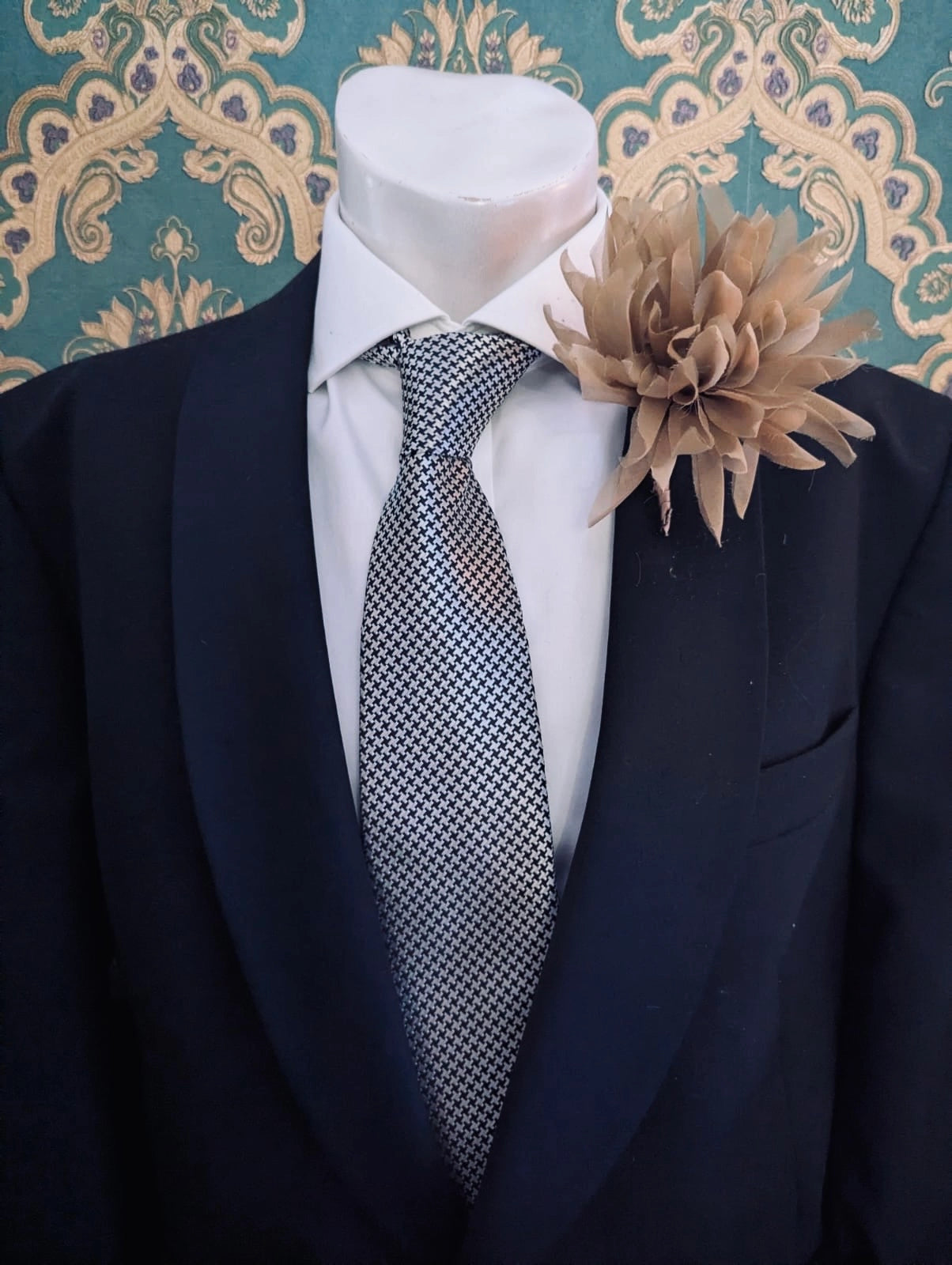 A black suit jacket displayed on a white mannequin torso, styled with a white dress shirt, a patterned grey and white tie, and a beige Broochella dahlia flower brooch against a teal and gold damask wallpaper background.