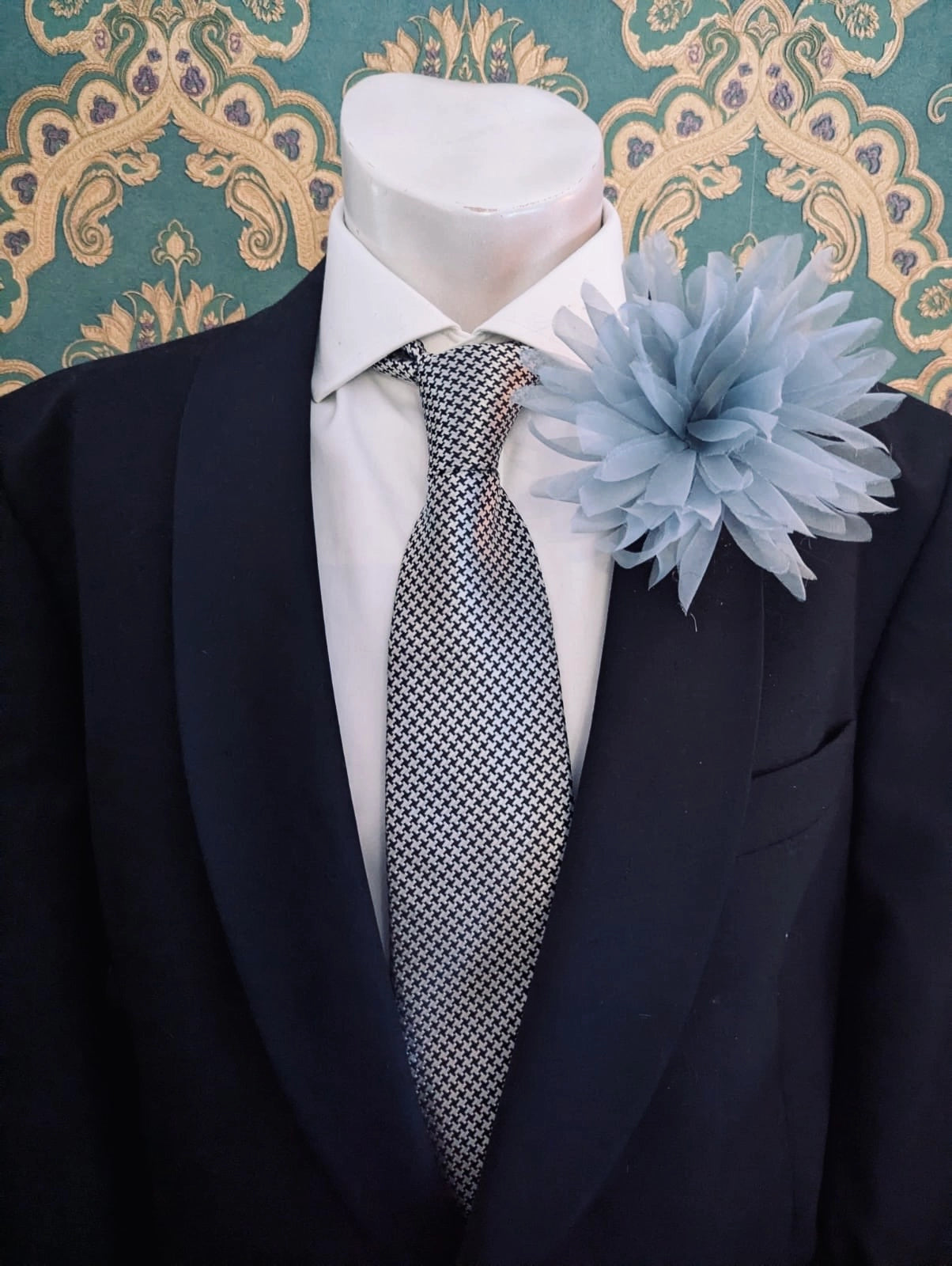 A black suit jacket displayed on a white mannequin torso, styled with a white dress shirt, a patterned grey and white tie, and a grey Broochella dahlia flower brooch against a teal and gold damask wallpaper background.