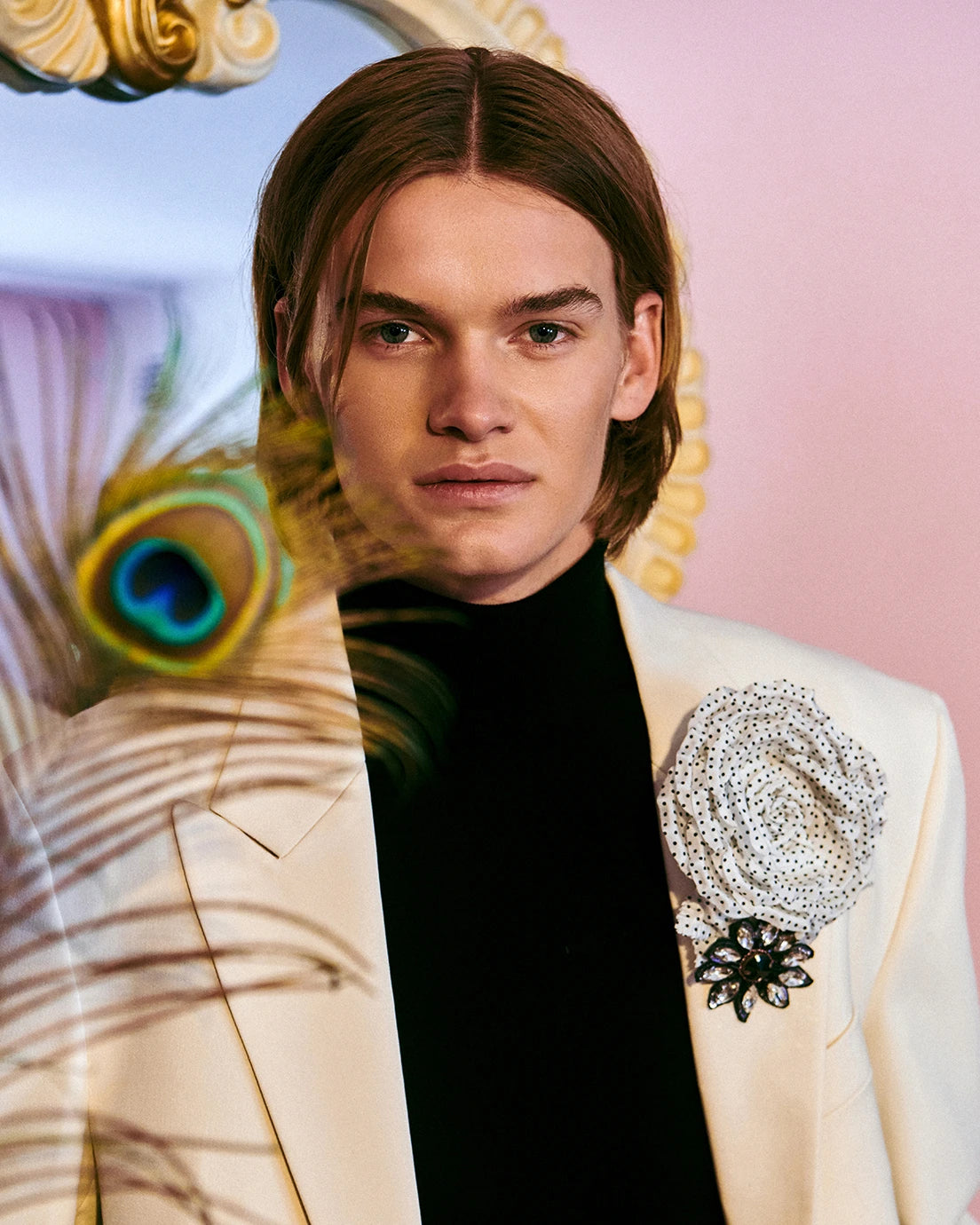 A man with long brown hair wearing a cream-colored blazer over a black turtleneck, featuring a large beaded floral lapel pin on the chest. He is wearing the Broochella men's Dalmata handmade flower brooch in white with black polka dots.  He is posed in front of a pink background with a gold mirror frame and a peacock feather overlay.