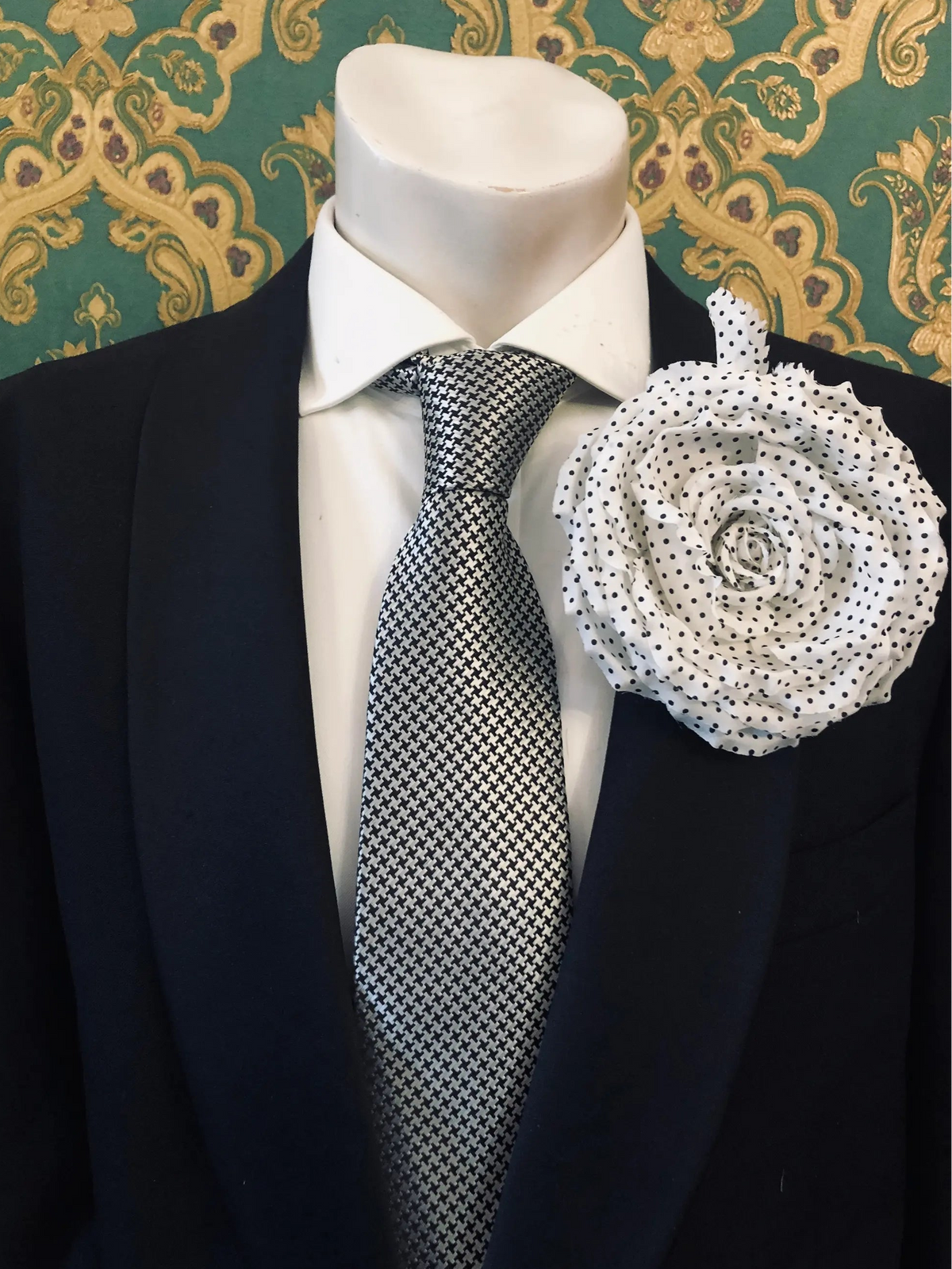 A Broochella Men's Dalmata white fabric flower brooch with black polka dots pinned to the lapel of a black suit jacket, styled with a white dress shirt and a black-and-white checkered tie, set against a green and gold patterned wallpaper background.