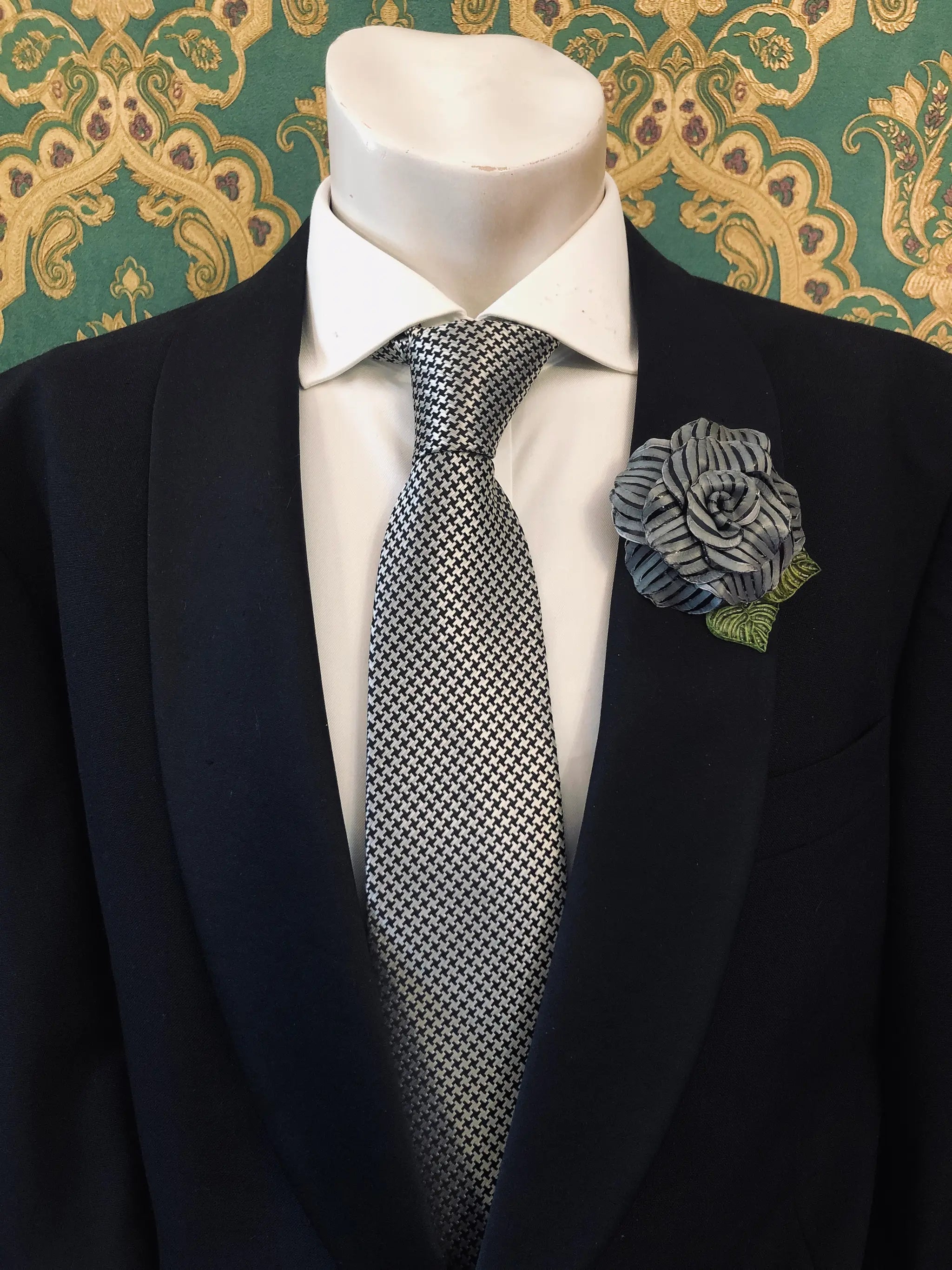 A men's formal outfit displayed on a white mannequin torso, featuring a black suit jacket, a white dress shirt, a grey and black checkered tie. Pinned on the lapel is a Broochella Men's Fabrice Camelia brooch in black with green leaves. The background is a green and gold patterned wallpaper.