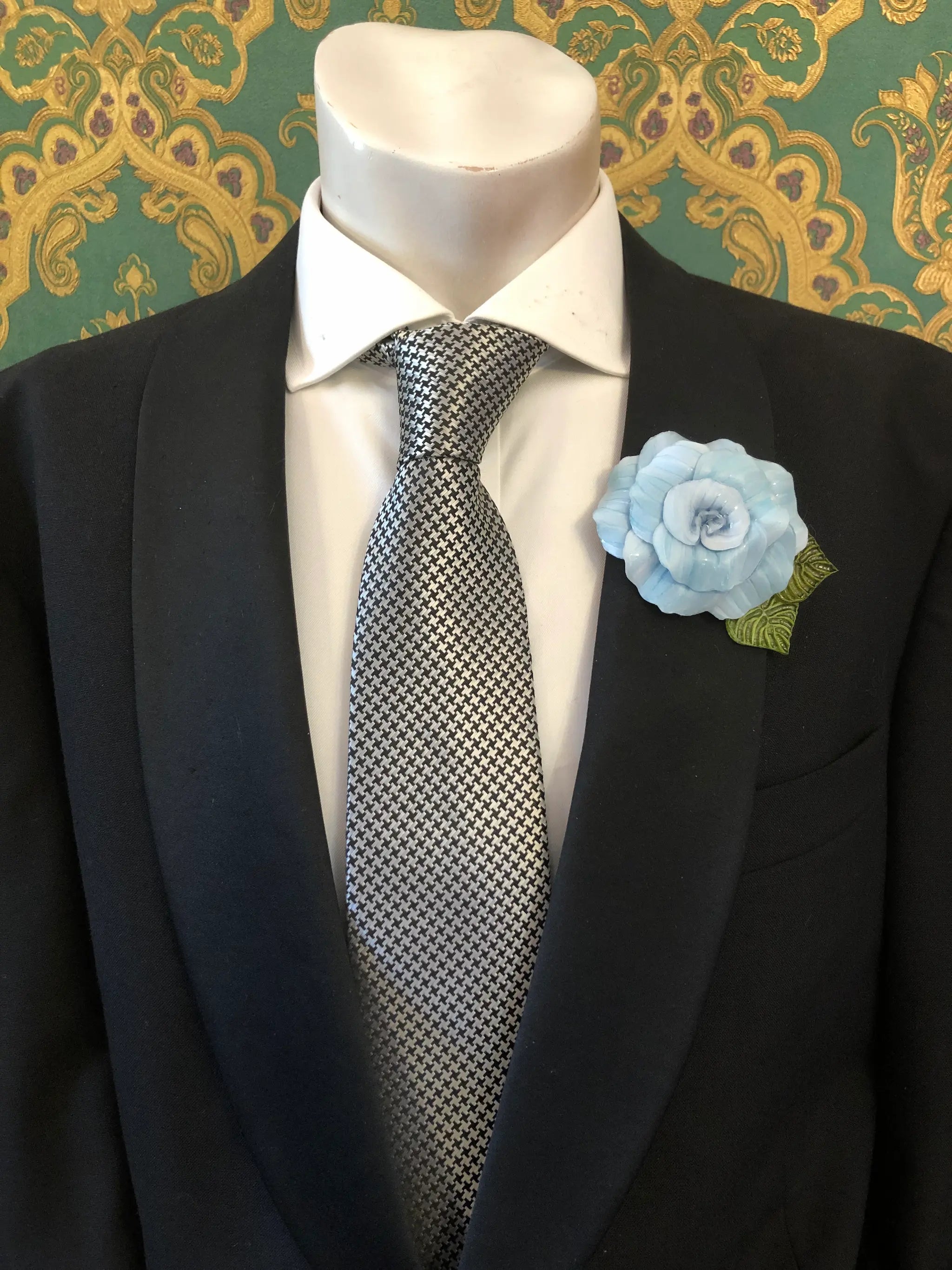 A men's formal outfit displayed on a white mannequin torso, featuring a black suit jacket, a white dress shirt, a grey and black checkered tie. Pinned on the lapel is a Broochella Men's Fabrice Camelia brooch in blue with green leaves. The background is a green and gold patterned wallpaper.