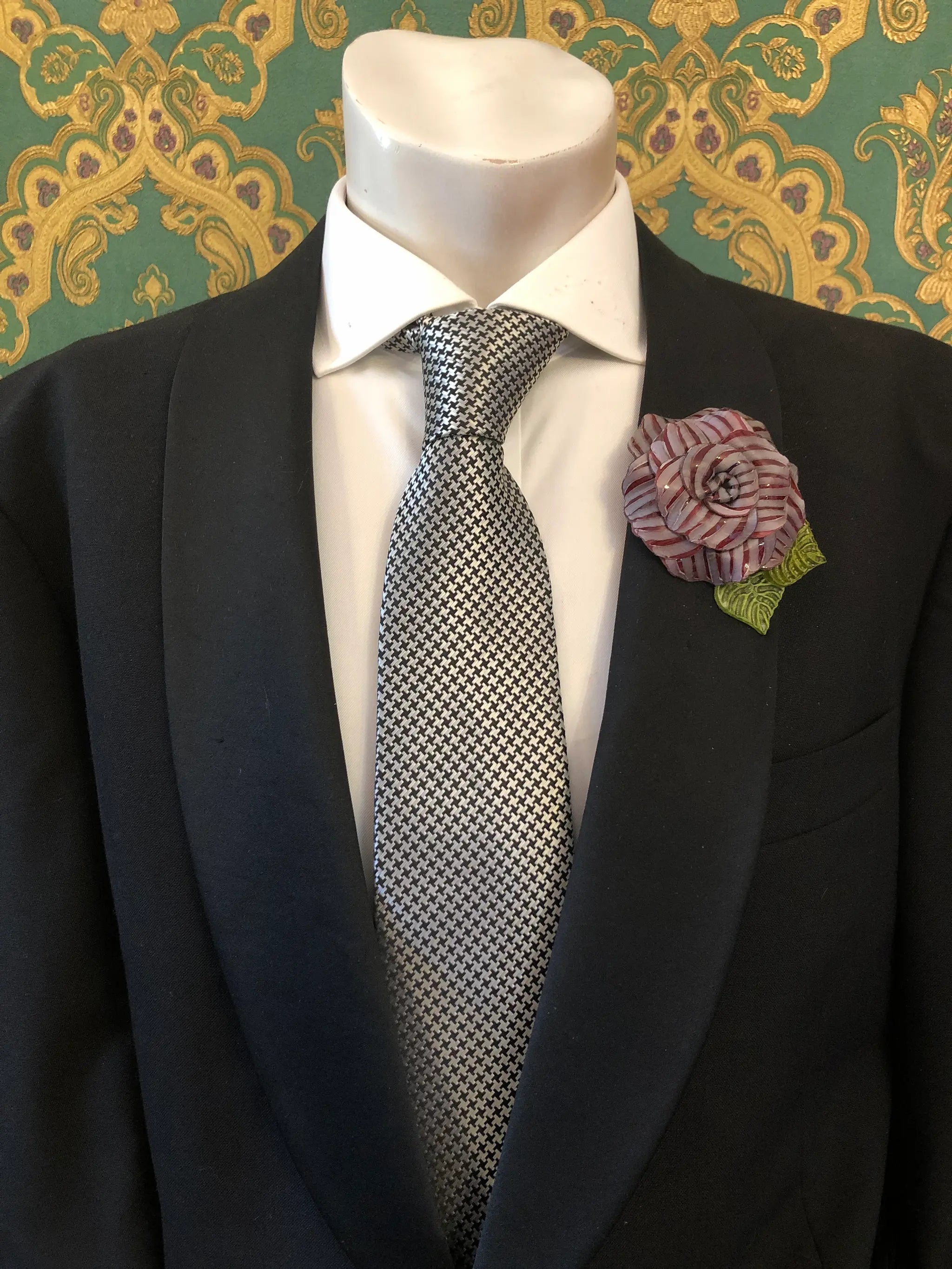 A men's formal outfit displayed on a white mannequin torso, featuring a black suit jacket, a white dress shirt, a grey and black checkered tie. Pinned on the lapel is a Broochella Men's Fabrice Camelia brooch in burgundy with green leaves. The background is a green and gold patterned wallpaper.